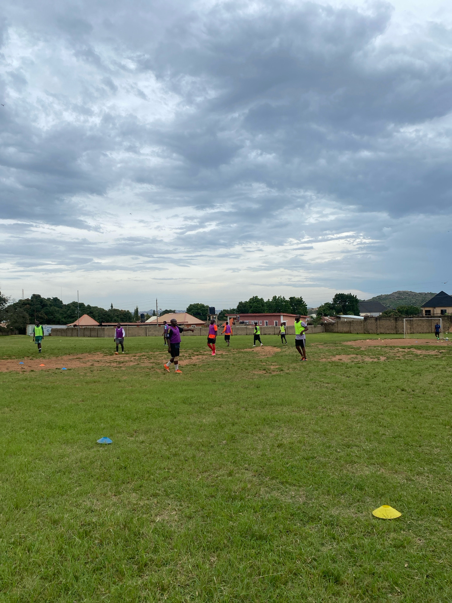 The image depicts an outdoor soccer practice session taking place on a grass field in Jos, Nigeria. The sky is overcast with dense, gray clouds. In the foreground, the grassy field is uneven and sparsely covered in some areas, with several orange and blue cones placed on the ground, indicating a training setup. In the middle ground, a group of men, likely athletes, are spread out across the field, engaged in various activities. Several individuals are wearing training bibs in different colors, such as yellow, purple, and orange. One person in the center of the frame, wearing a purple bib, is facing away from the camera and appears to be pointing or gesturing. Another individual in a green bib is further to the left, and others are scattered throughout the area, some in motion. In the background, there is a low stone wall enclosing the field, with residential buildings and trees visible beyond it. To the far right, a soccer goal is set up, and one person is near it. The overall scene suggests a casual training session under a cloudy sky.
