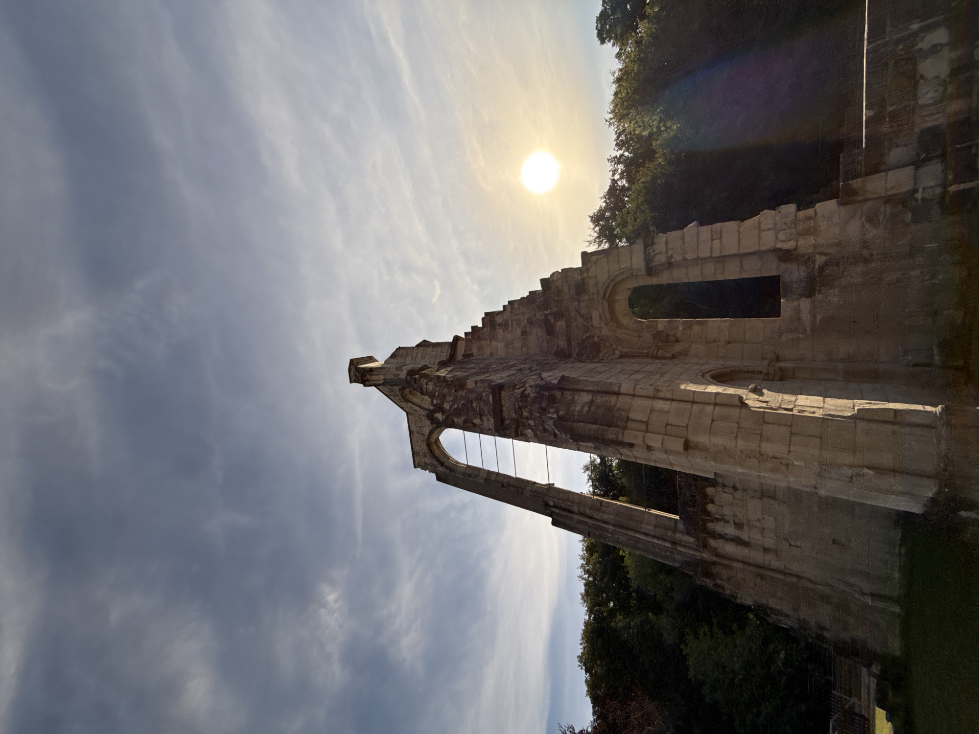 An outdoor, low-angle, wide shot captures the ruins of Walkenried Abbey in Walkenried, Germany. The sky is filled with a mix of blue and gray clouds, with the sun appearing as a bright, yellow orb in the upper right quadrant, casting a lens flare downwards. The sun is partially obscured by the sky, but its light illuminates the weathered, stone structure of the abbey in the foreground. The ruins are made of large, rough-hewn stones, with gaping arched windows and a tall, pointed facade suggesting a former Gothic architectural style. Green foliage and trees form a dense line behind the abbey ruins, creating a contrast between the man-made structure and the natural environment. The ground in front of the abbey is covered with short, green grass. The overall impression is one of historical decay and natural reclamation.