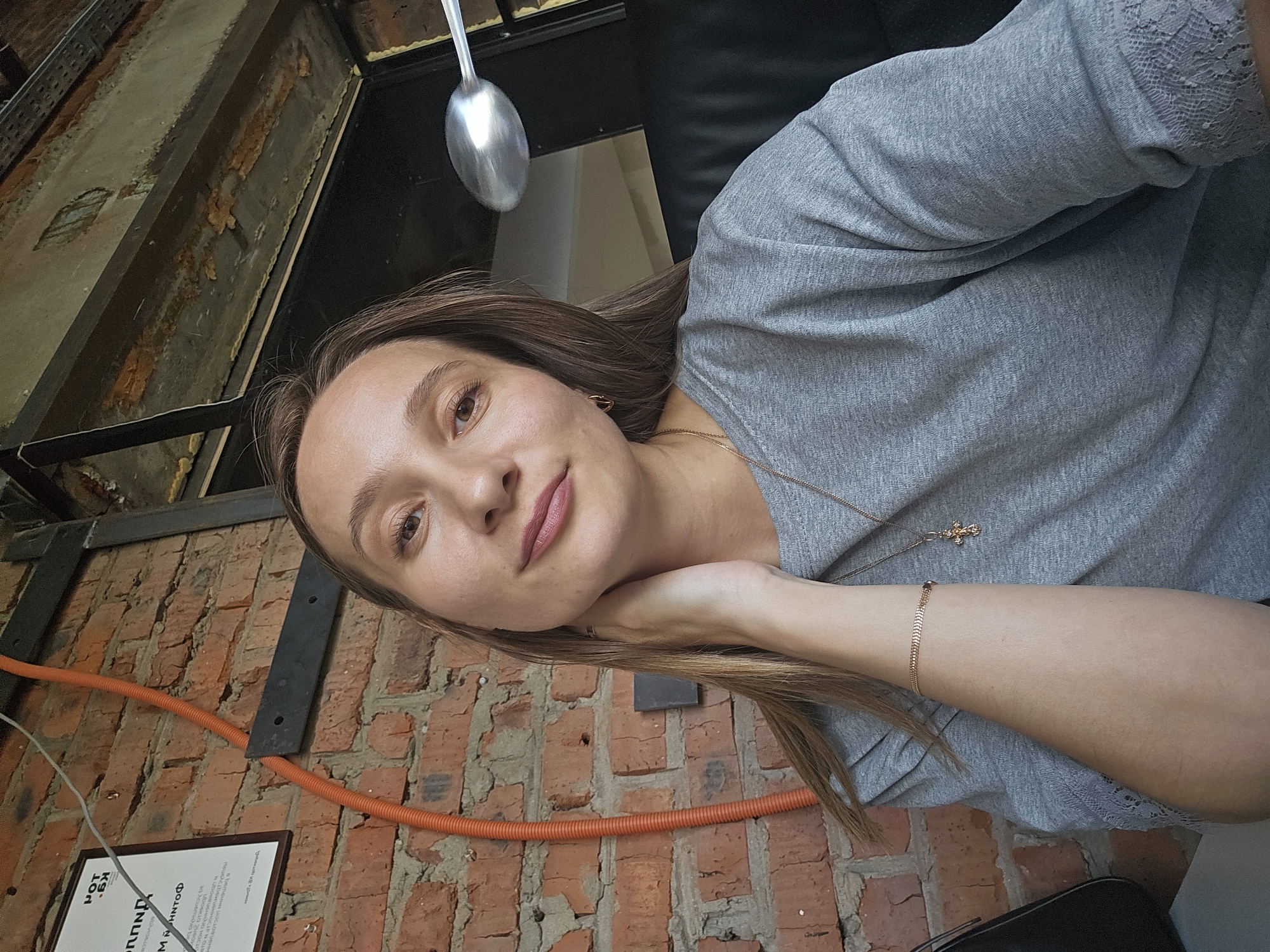 A young woman is featured in a close-up, slightly tilted portrait, looking directly at the camera with a gentle, content expression. She has brown eyes, fair skin, and long brown hair. Her left hand is positioned under her chin, with her fingers resting on her neck. She wears a light gray, long-sleeved top with a subtle texture and lace detailing on the sleeve, a delicate gold-colored necklace with a small cross pendant, and a matching chain bracelet on her left wrist.
The background reveals an indoor setting with an industrial-rustic aesthetic. A prominent exposed brick wall is visible, with an orange corrugated electrical conduit running horizontally across it. Above the brick wall, metal beams and structural elements suggest exposed ceiling or wall components, possibly with insulation or unfinished surfaces. A large, reflective silver spoon is visible in the upper center of the frame, positioned above her head.
In the lower left corner, a framed document or sign is partially visible on the brick wall. Recognizable Cyrillic text includes "POT" and "ИМЯ НИНТОФ". The scene suggests a relaxed atmosphere, possibly in a cafe, restaurant, or a repurposed industrial space in Chaikovsky, Russia. The lighting is ambient, typical of an indoor environment.