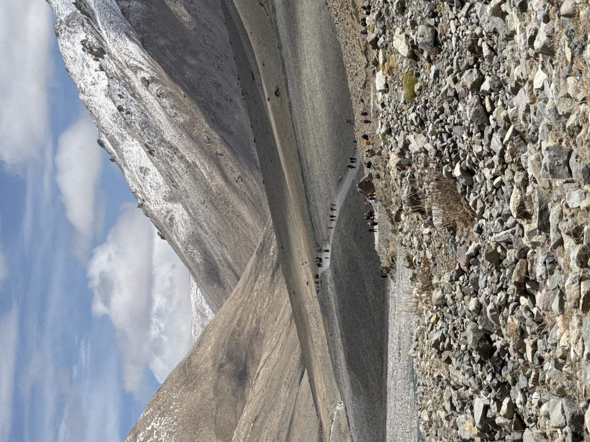 The image depicts a mountain landscape, likely in a remote region. A line of people is walking along a narrow path through the rocky terrain. The environment is arid and mountainous, with a partly cloudy sky visible. There is no indication of this image being in Parga town, China. It seems more like a mountainous region in a different location.