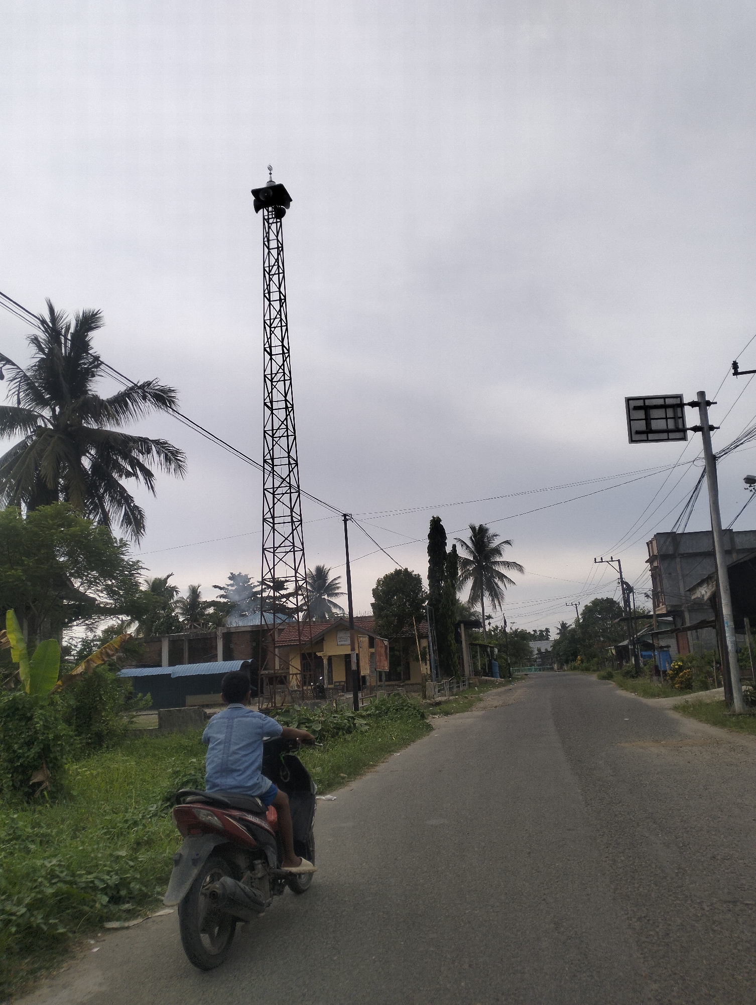 A young boy in a blue shirt and shorts rides a red and black scooter on a paved road. He is facing away from the camera. The road is lined with green vegetation and some buildings. In the background, a tall, black lattice tower stands against a cloudy sky. The tower has speakers at the top. Further down the road, more buildings and trees are visible. The scene appears to be in a rural or semi-urban area in Indonesia, possibly Kilometer Delapan, given the context. The weather is overcast, suggesting it might be morning or late afternoon. There are no visible signs of text in the image.