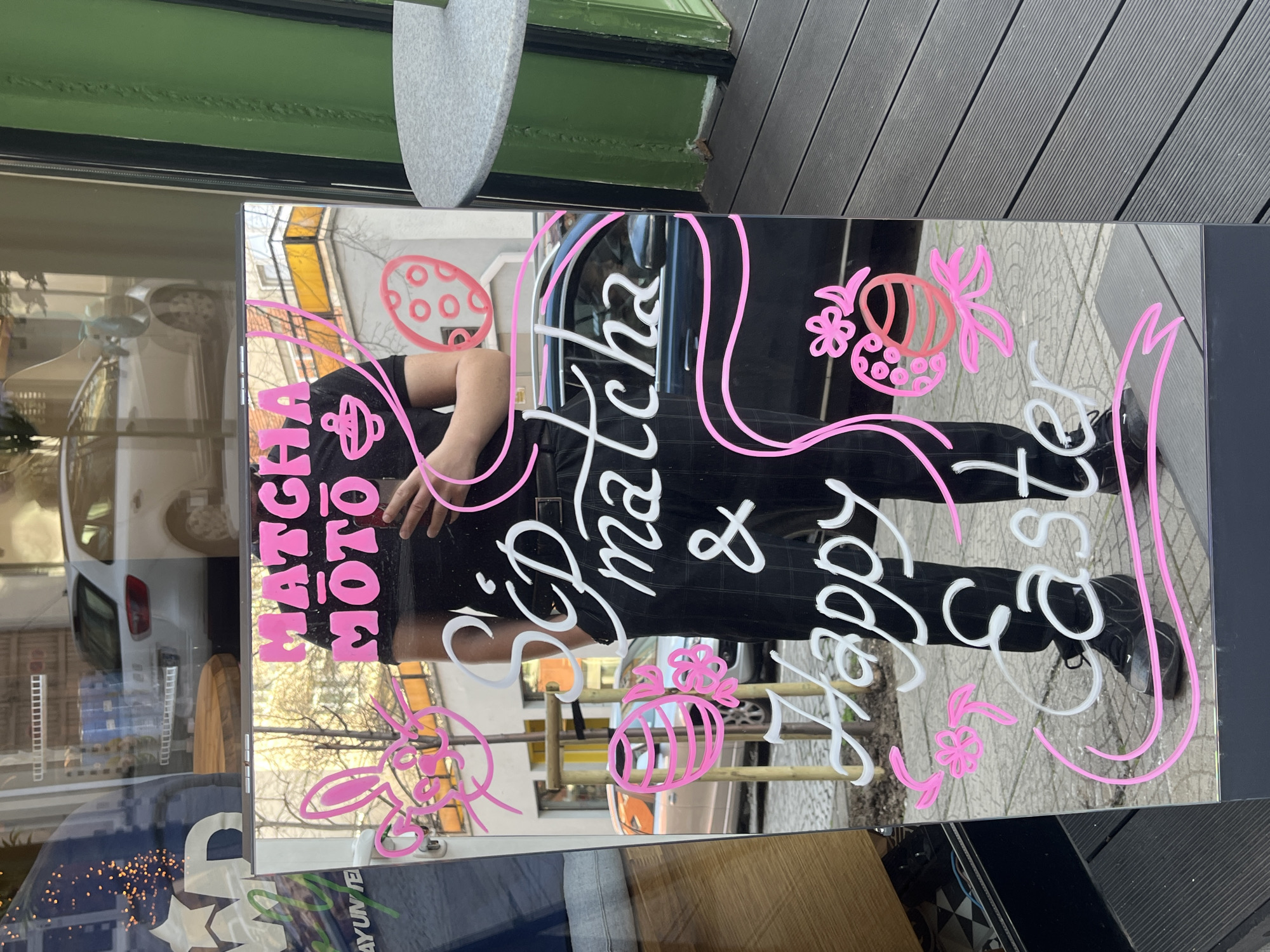 The image shows a mirrored sandwich board sign in front of a business in Sofia, Bulgaria. The sign advertises "Matcha MOTO & Happy Easter" written in pink and white paint, adorned with Easter egg and flower decorations. The reflection in the sign reveals a person standing behind it, wearing a black t-shirt and checkered pants. The background shows a street scene with buildings, trees, and parked cars. The sign is positioned on a sidewalk near a green pillar and a wooden deck.