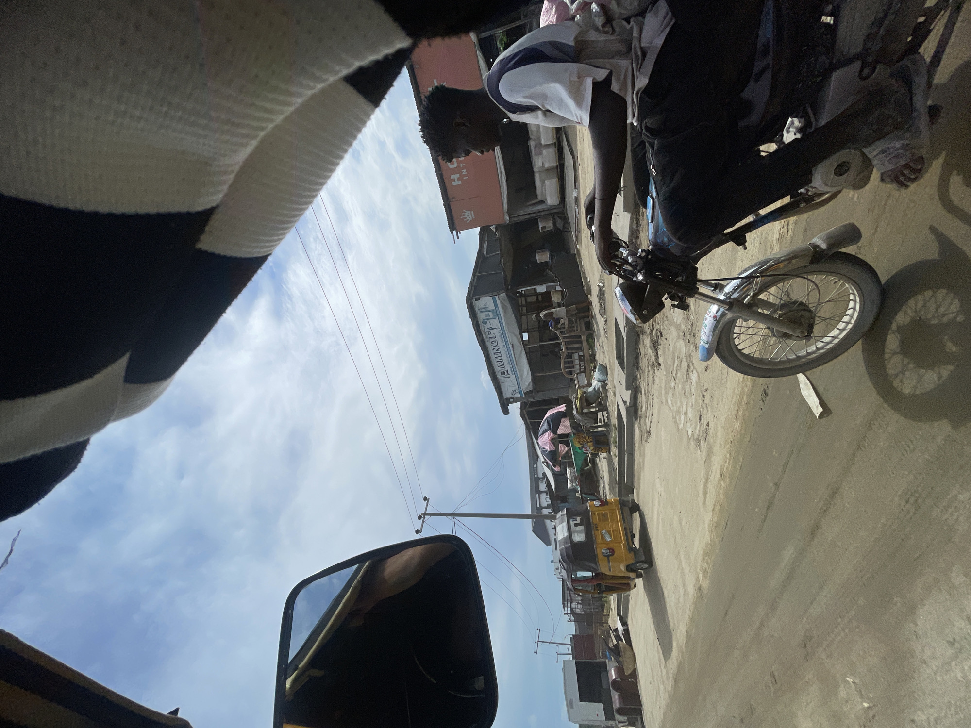 A street scene in Ado, Nigeria, shows a man on a motorcycle, buildings lining the road, and a yellow tricycle (Keke) in the distance. Power lines stretch across the sky. The view is partially obstructed by a black and white striped object and a car mirror.