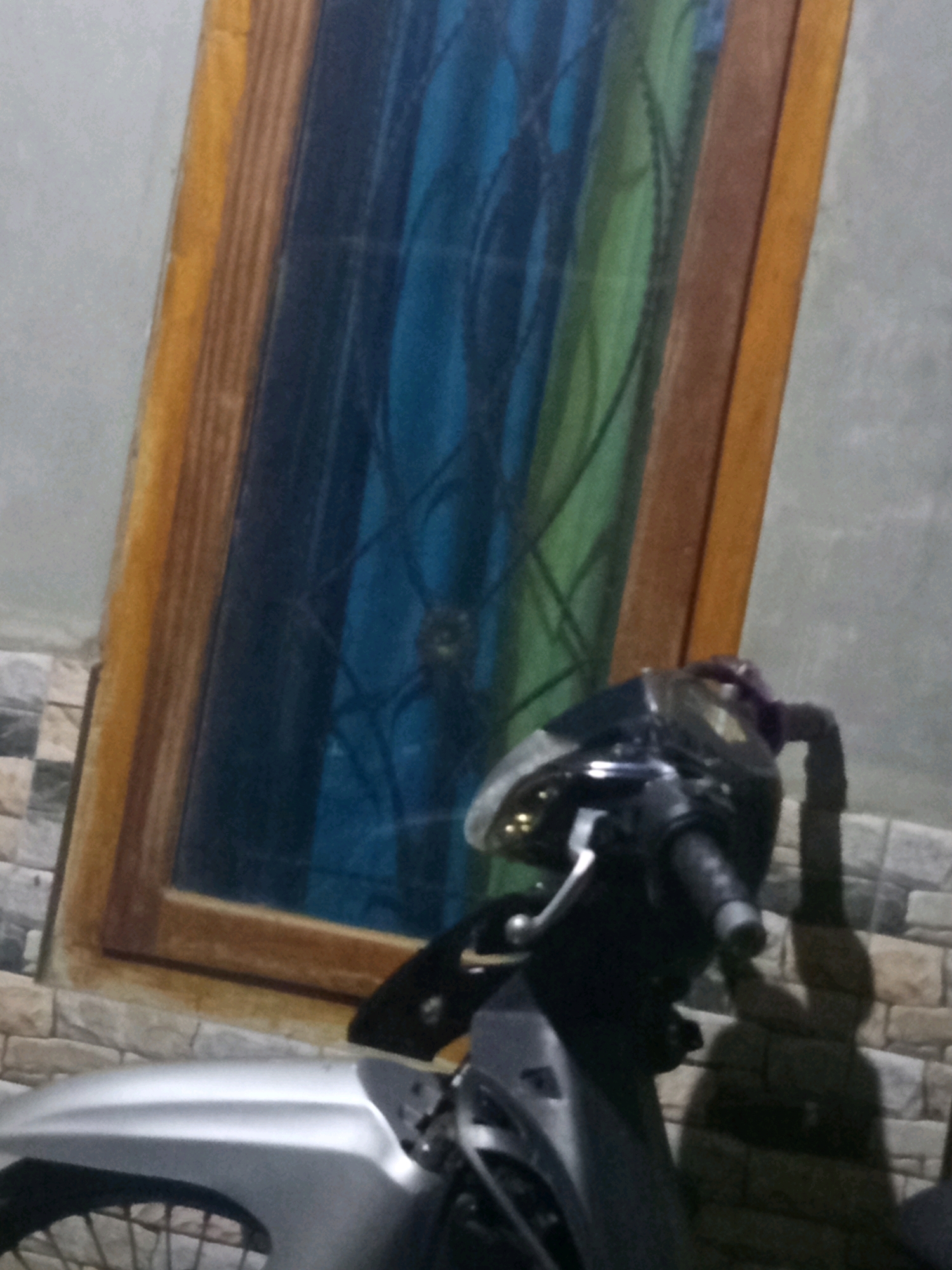 The image shows a close-up view of a motorcycle parked in front of a building. The motorcycle is silver and black and is positioned on the right side of the frame. A window with blue and green curtains is visible in the background, framed by a wooden border. The wall behind the window is gray, and the lower part of the wall is covered with brick tiles. A shadow of a person is cast on the brick tiled wall to the right of the motorcycle. The scene appears to be an outdoor area in front of a house in Karang Sari, Indonesia.