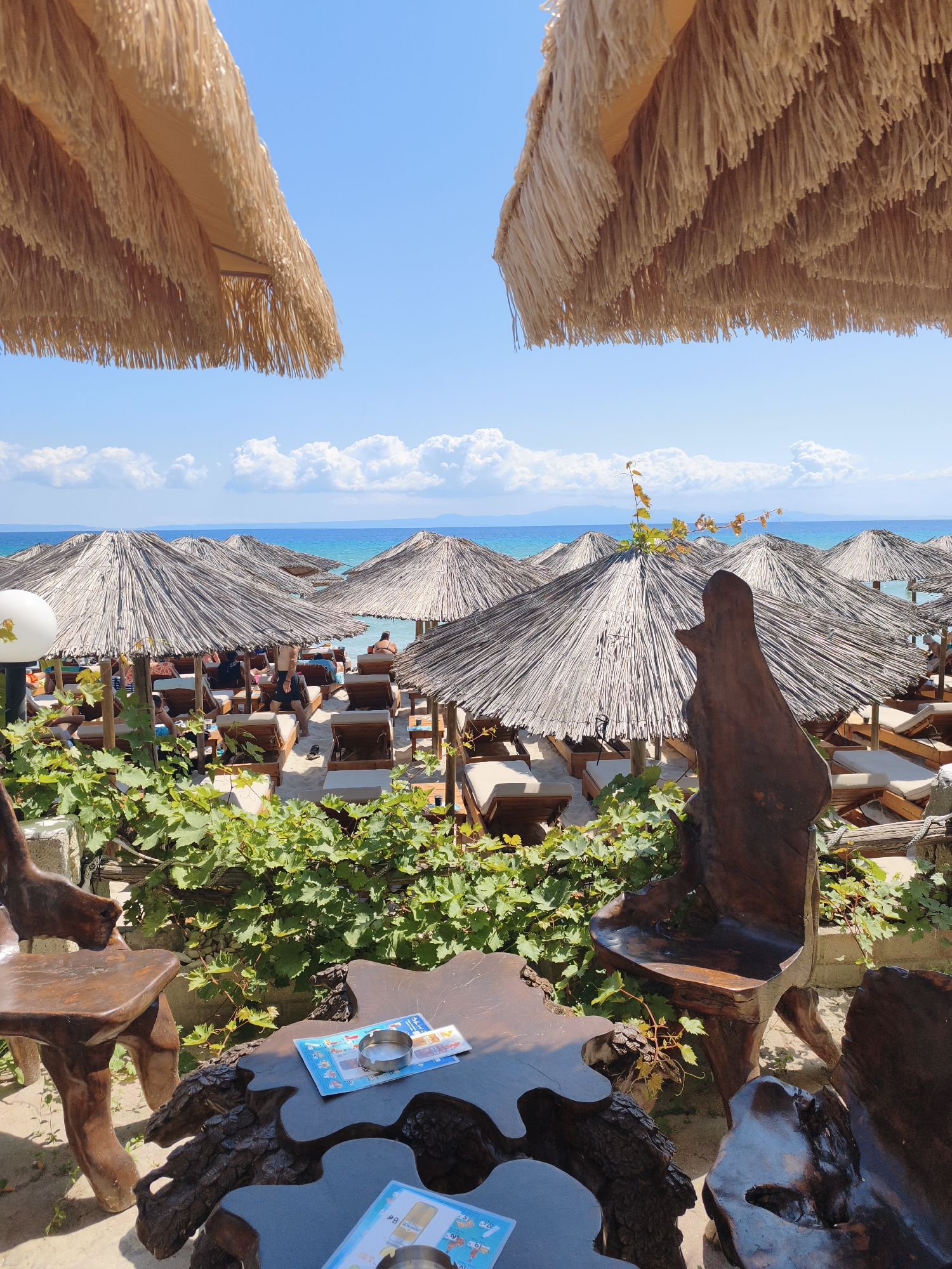 The image depicts a beach resort scene in Kassandra Municipal Unit, Greece, on a clear day with a blue sky and scattered clouds. In the foreground, two rustic, carved wooden chairs flank a similarly crafted wooden table. On the table, two menus and an ashtray are visible. The table appears to be made from tree trunks and roots, with an irregular, organic shape.
Beyond the seating area, a beach is populated with numerous thatched-roof umbrellas providing shade for rows of lounge chairs. Some people are seated on the lounge chairs, appearing relaxed. The sand is light-colored, and the turquoise ocean stretches to the horizon, with distant landmasses visible. Green foliage, likely from vines, partially obscures the view of the beach setup from the front. Two large thatched umbrellas, framing the top of the image, are prominently displayed, extending into the sky. The overall atmosphere suggests a tranquil and inviting beachside establishment.