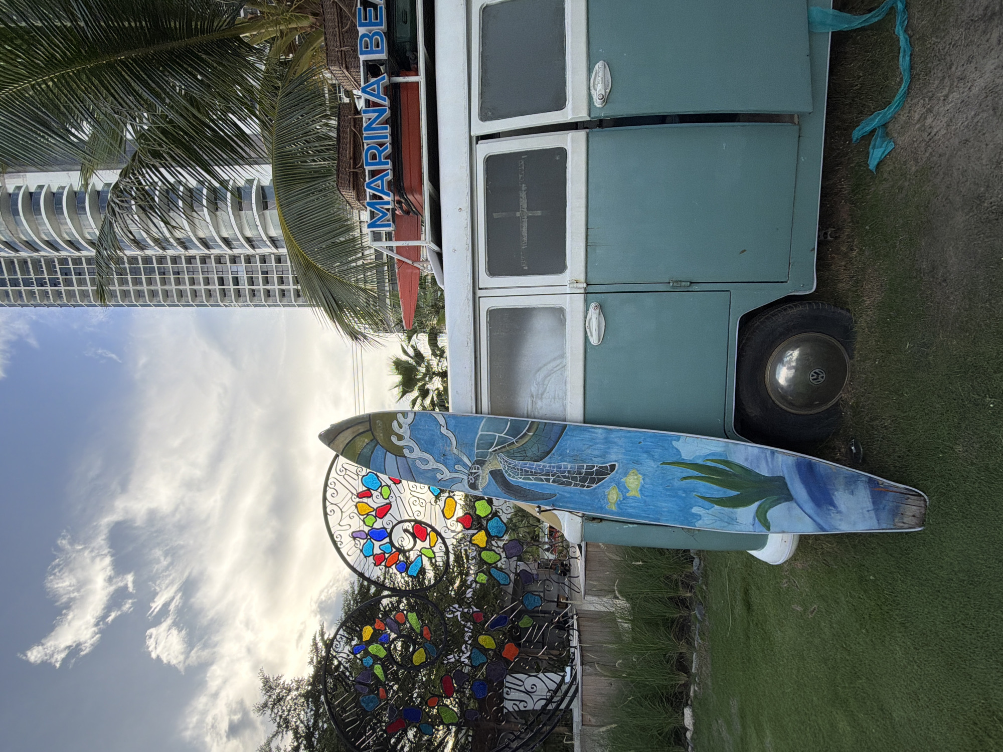 An old turquoise and white Volkswagen van is parked on a grassy area. A surfboard with a painted sea turtle and fish design is propped up against the side of the van. In the background, a modern high-rise building stands tall against a cloudy sky. To the left of the van, there is an intricate metal sculpture with colorful glass inserts. The overall scene appears to be a roadside attraction or a scenic viewpoint. It is likely located in the city of Nha Trang, Vietnam, known for its beaches and coastal attractions.