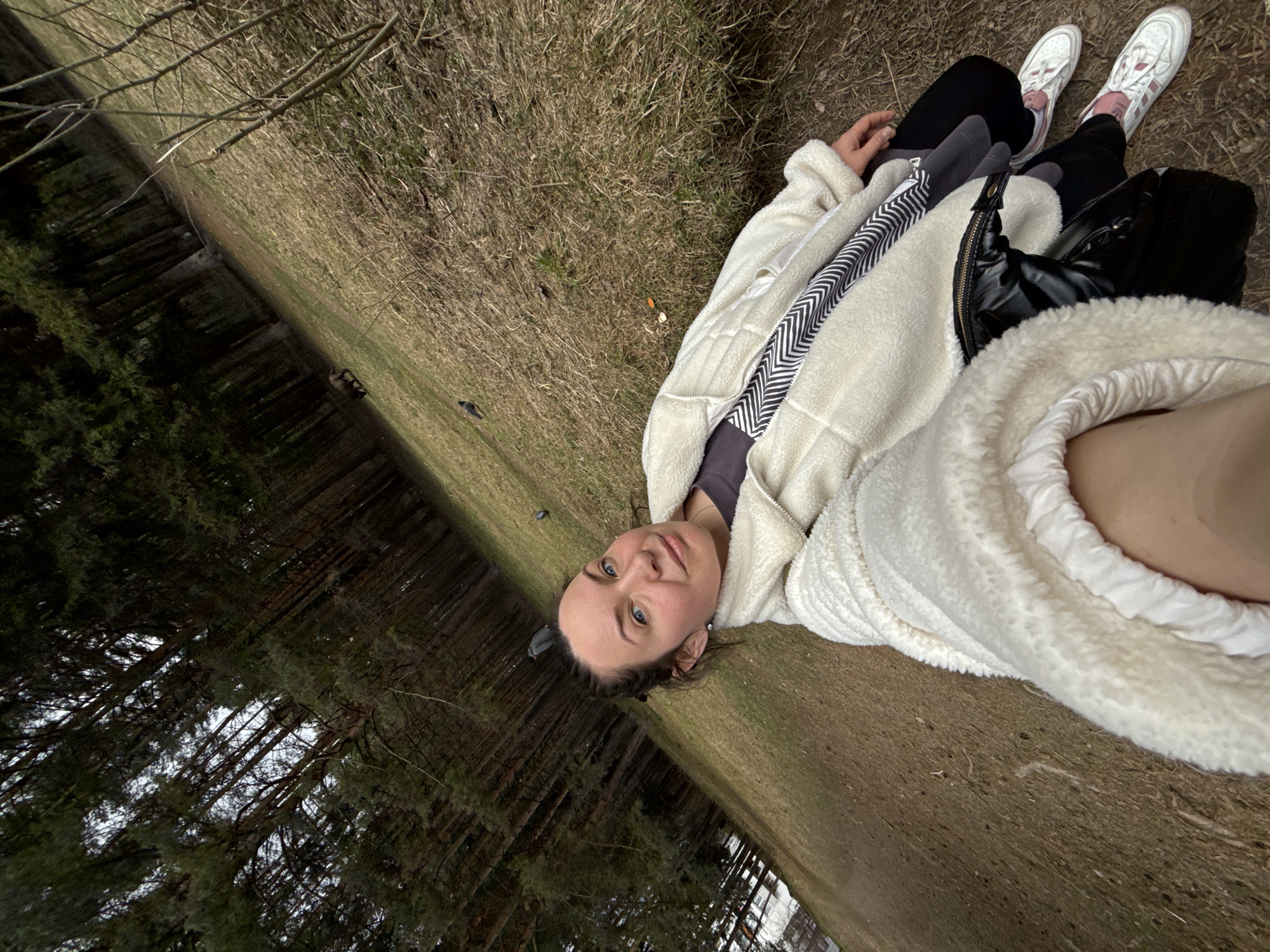 A young woman is depicted lying on dry grass at the edge of a forest, looking towards the camera with a calm and pleasant expression. Her dark hair is pulled back, and she has light blue eyes and fair skin. She is wearing a cream-colored, fluffy jacket, possibly made of sherpa or fleece, over a dark top. A black and white chevron or striped pattern is visible on the inner lining of her jacket. She also wears black pants and white athletic shoes with pink accents. A black handbag or backpack is seen beside her feet.
The scene is an outdoor natural environment, likely a park or the outskirts of a forest in Chaikovsky, Russia. The ground is covered in dormant, light brown grass, suggesting late winter or early spring. In the background, there is a dense stand of tall, dark evergreen trees, possibly pines or firs, with their trunks forming a dark, vertical pattern against the lighter sky visible through the canopy. Further back on the grass, two dark birds are visible, one appearing to be a duck and the other a smaller, darker bird. The lighting is soft, indicative of an overcast day or indirect sunlight. The image is tilted, providing a dynamic perspective. No visible text is present.