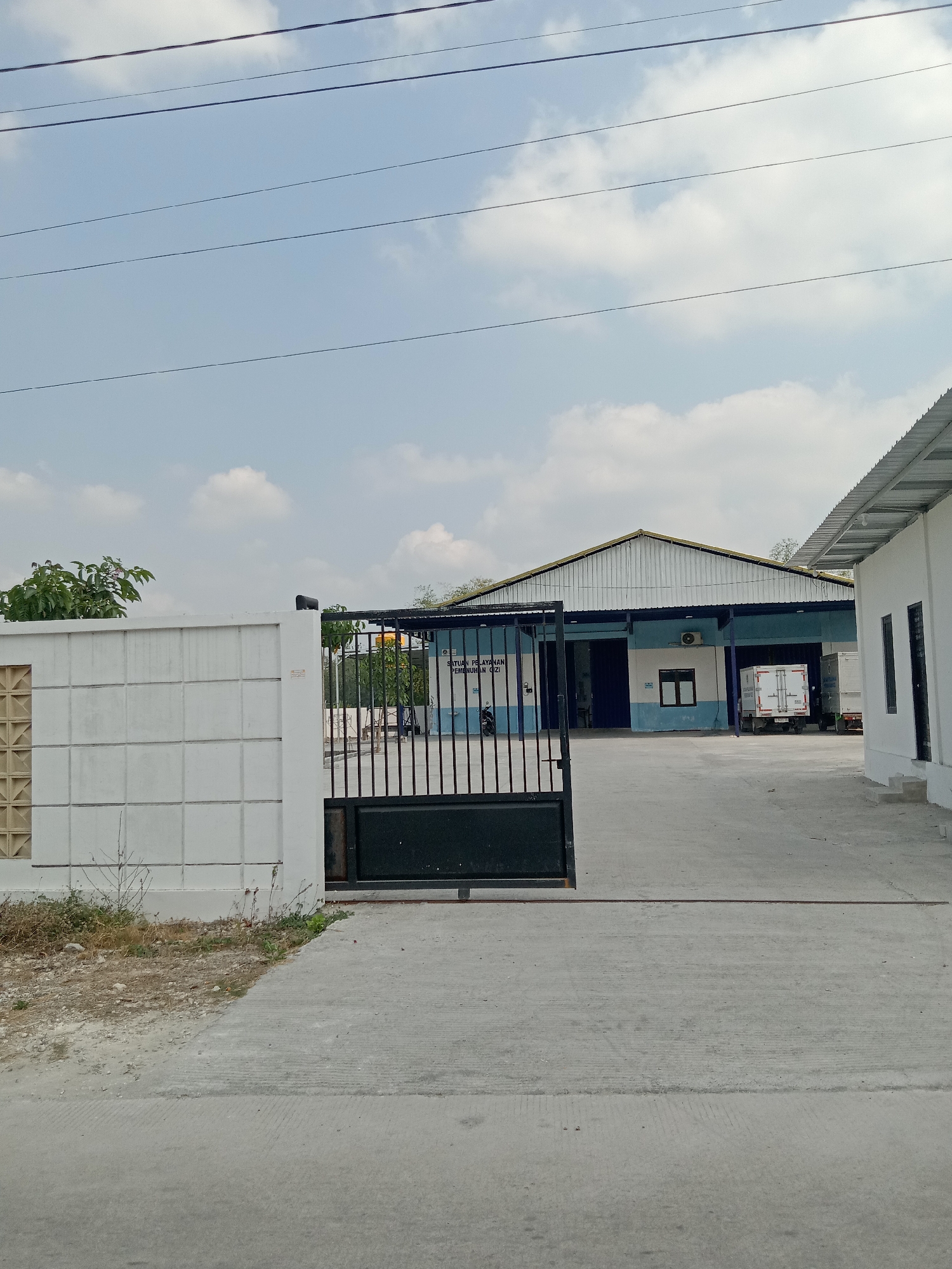 This is a photograph taken on a cloudy day in Sidobandung, Indonesia, showing a street view of what appears to be an industrial or commercial facility. In the foreground, there is a concrete road leading to a black metal gate. To the left of the gate is a white concrete wall with square indentations. Through the gate, a large building with a blue and white facade and a corrugated metal roof is visible. The building has large open bays, one of which contains a white truck with a refrigeration unit. To the right of the main building, another white building with a corrugated metal roof is partially visible. The sky is filled with scattered white clouds. The overall scene is industrial, with elements of infrastructure and logistics evident.
