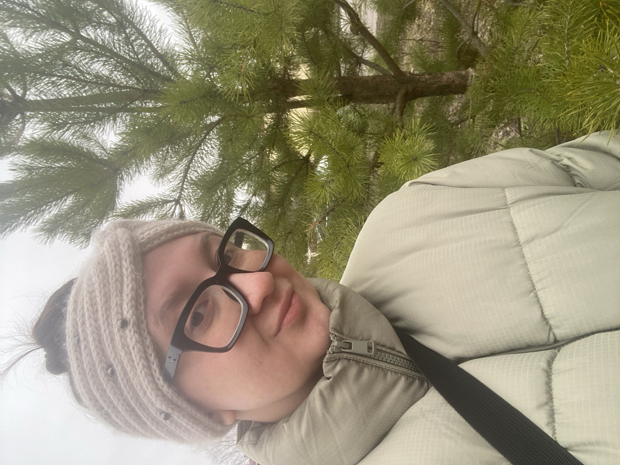 A selfie of a woman with light skin is taken in front of a pine tree. She wears a light-colored knit headband with small metal embellishments, black-framed glasses, and a light green puffer jacket with a black strap across her chest. The background is a blurred view of the pine tree, suggesting an outdoor setting, possibly a park or forest area. The location may be in Chaikovsky, Russia.