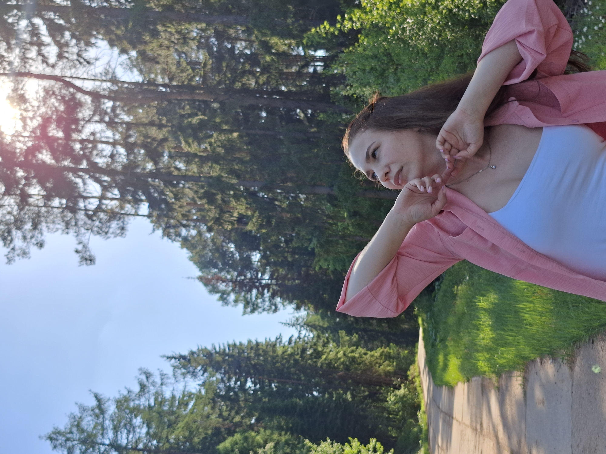 A young woman stands on a paved path in Прикамский, Russia. She wears a white top and a pink jacket. She has long brown hair and is touching her face with her hands, looking to the left. Behind her are tall trees with green leaves, partially obscuring a blue sky with the sun shining through. The path is lined with green grass.