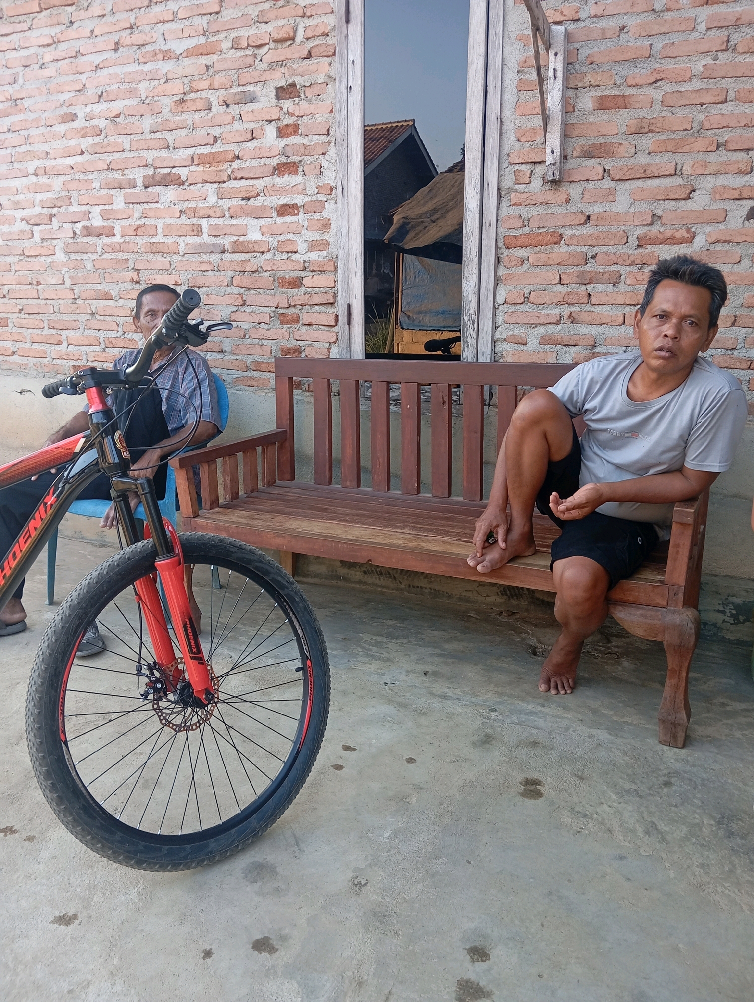 Two men are present in the image. The man on the left is seated on a blue plastic chair, leaning back. A red and black bicycle is positioned in front of him, with its front wheel prominently in the foreground. He is wearing a checkered shirt and dark pants. The man on the right is seated on a wooden bench, with his legs crossed. He is wearing a light gray t-shirt and black shorts. Both men appear to be in their middle age.
The environment consists of a brick wall behind the men, a wooden bench, and a concrete floor. A window with a reflection of a structure with a corrugated roof is visible on the wall. The overall scene suggests an outdoor setting in a residential area. The image was taken in Adi Puro, Indonesia.