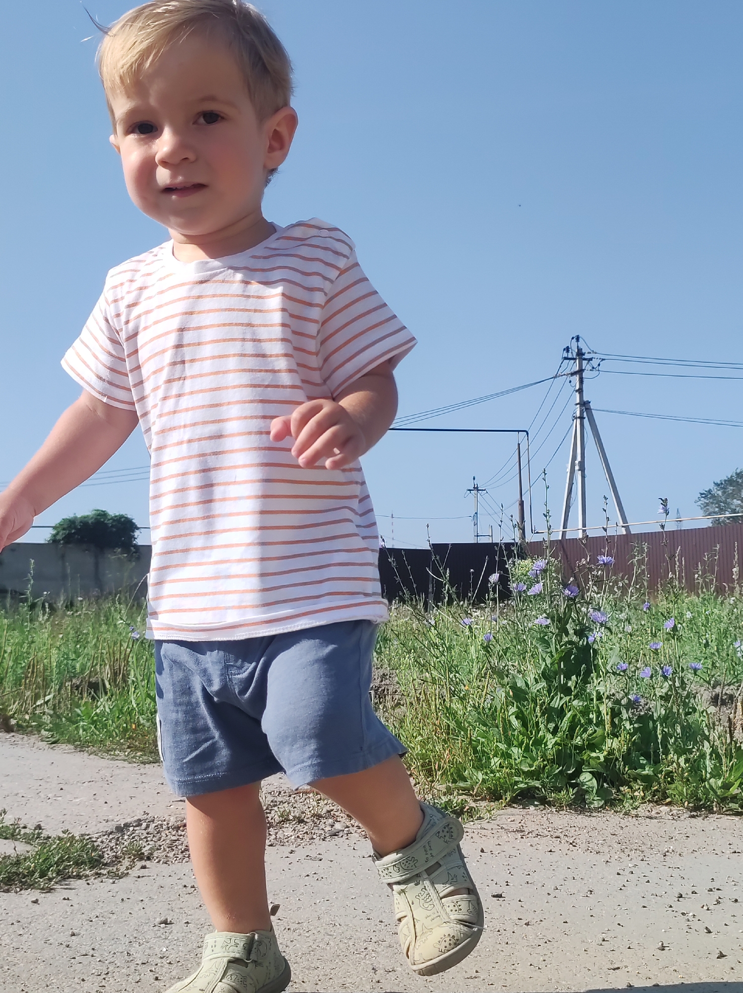 The image captures a young child with blonde hair walking outdoors on a sunny day. The child is wearing a horizontally striped t-shirt with orange and white stripes, blue shorts, and light green sandals with a dinosaur pattern. The child is looking slightly to the left and appears to be mid-stride. In the background, there is a clear blue sky, some green vegetation with small purple flowers, a wooden fence, and utility poles with wires. The ground is a dirt or gravel path. While the specific location within Chaikovsky, Russia, cannot be determined from the image alone, it depicts a common outdoor setting that could be found in a residential or rural area of the city.