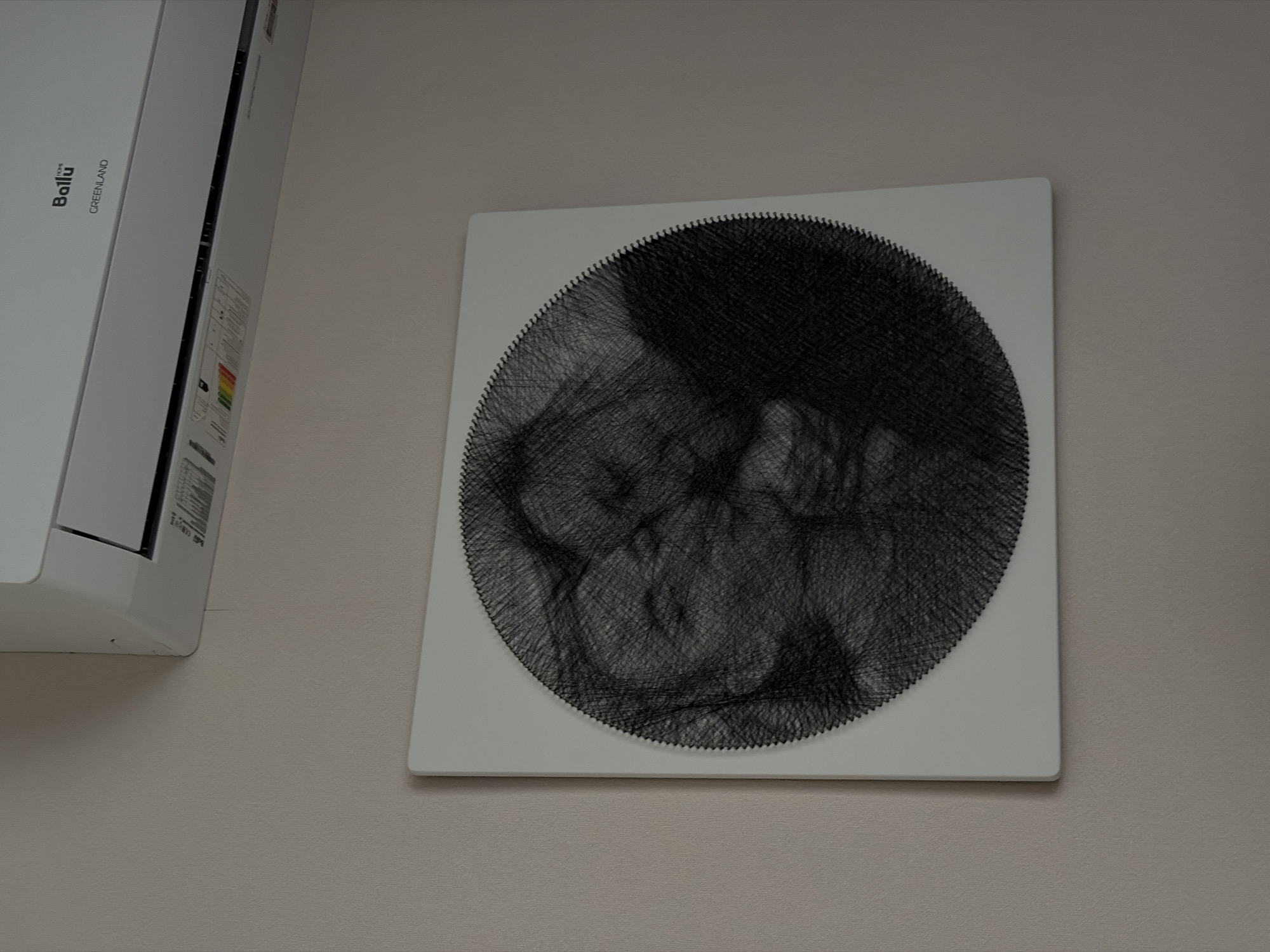 A white wall displays an air conditioner unit with "Ballu GREENLAND" printed on it and a square artwork featuring a black string art design of a sleeping baby in Krasnodar, Russia.
