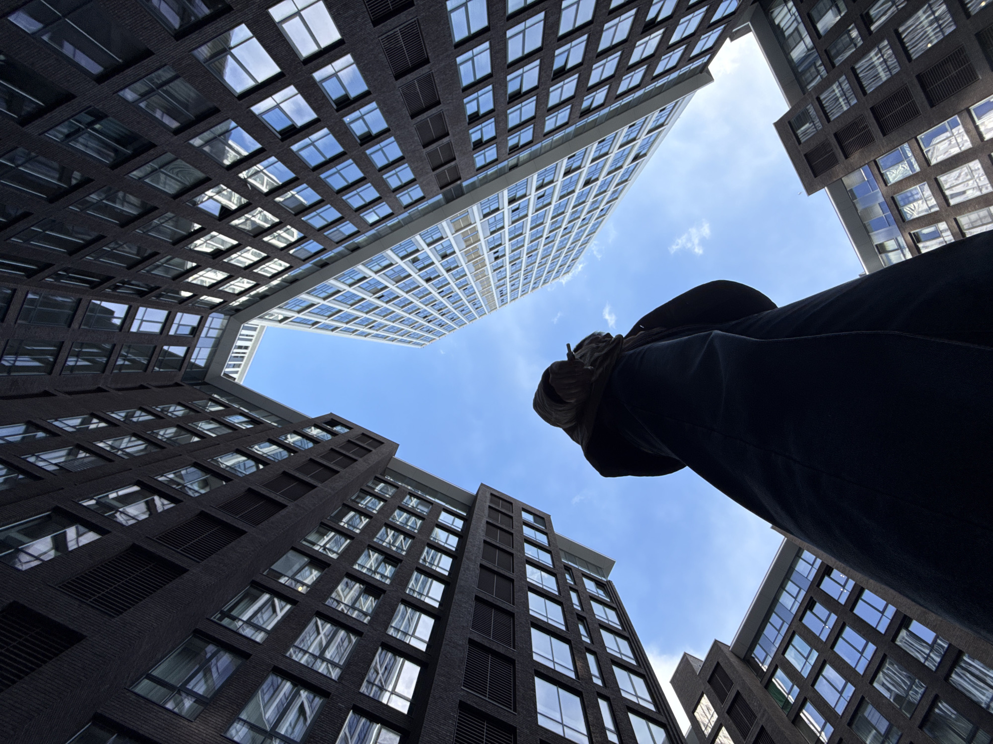 A low-angle perspective captures modern architecture against a blue sky with scattered clouds. Several buildings with geometric window patterns frame the view. A person's legs and torso intrude into the frame from the right, suggesting a first-person perspective looking up at the buildings. The buildings appear to be located in Moscow, Russia.