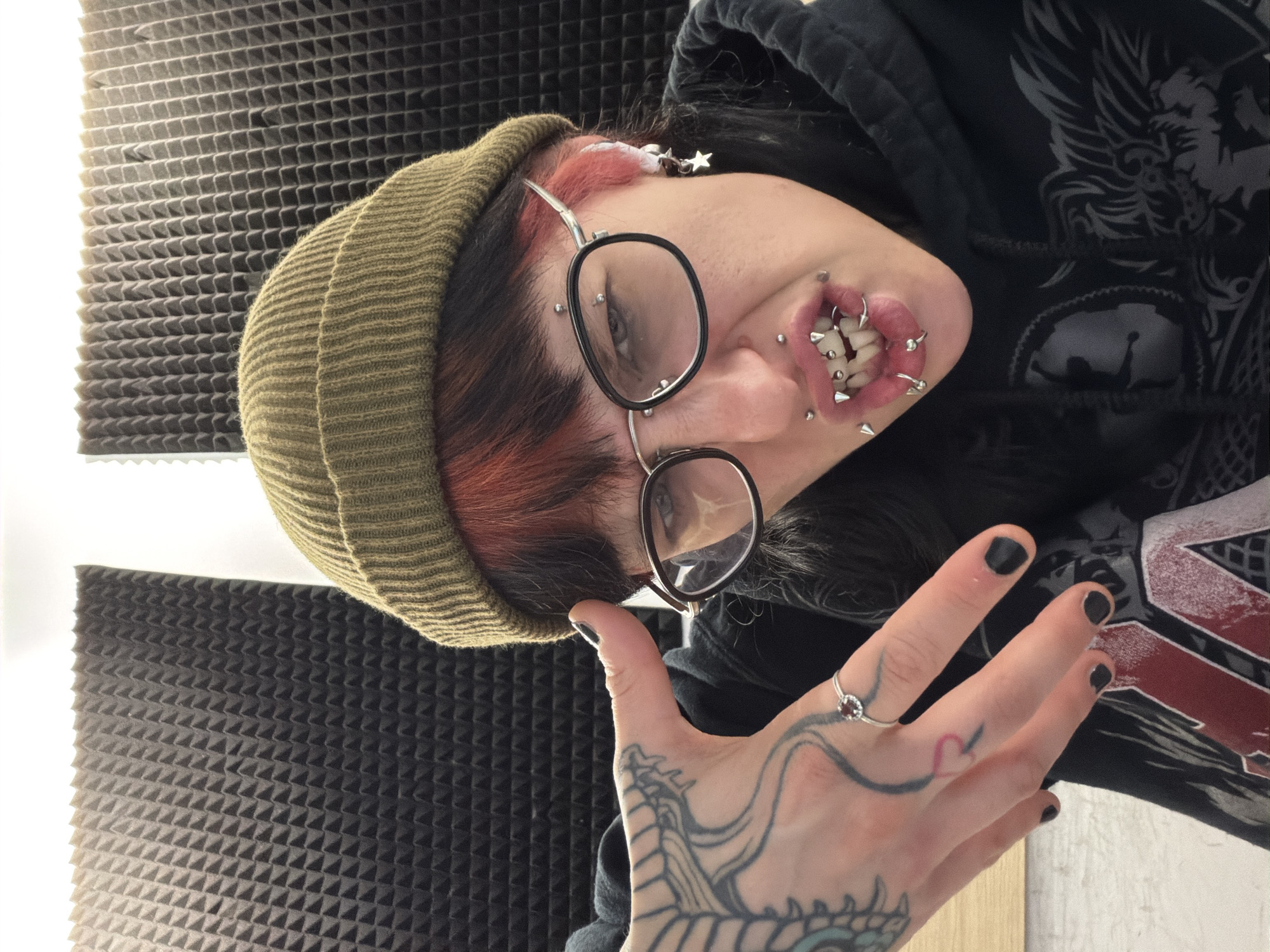 The image shows a person with multiple facial and lip piercings, wearing a green beanie and glasses, and a black hooded sweatshirt with a graphic design. The person has tattoos on their hand and black nail polish. The background is a wall covered in soundproofing foam. It's not possible to identify a location within the city of Sofia, Bulgaria, from the visual information provided.