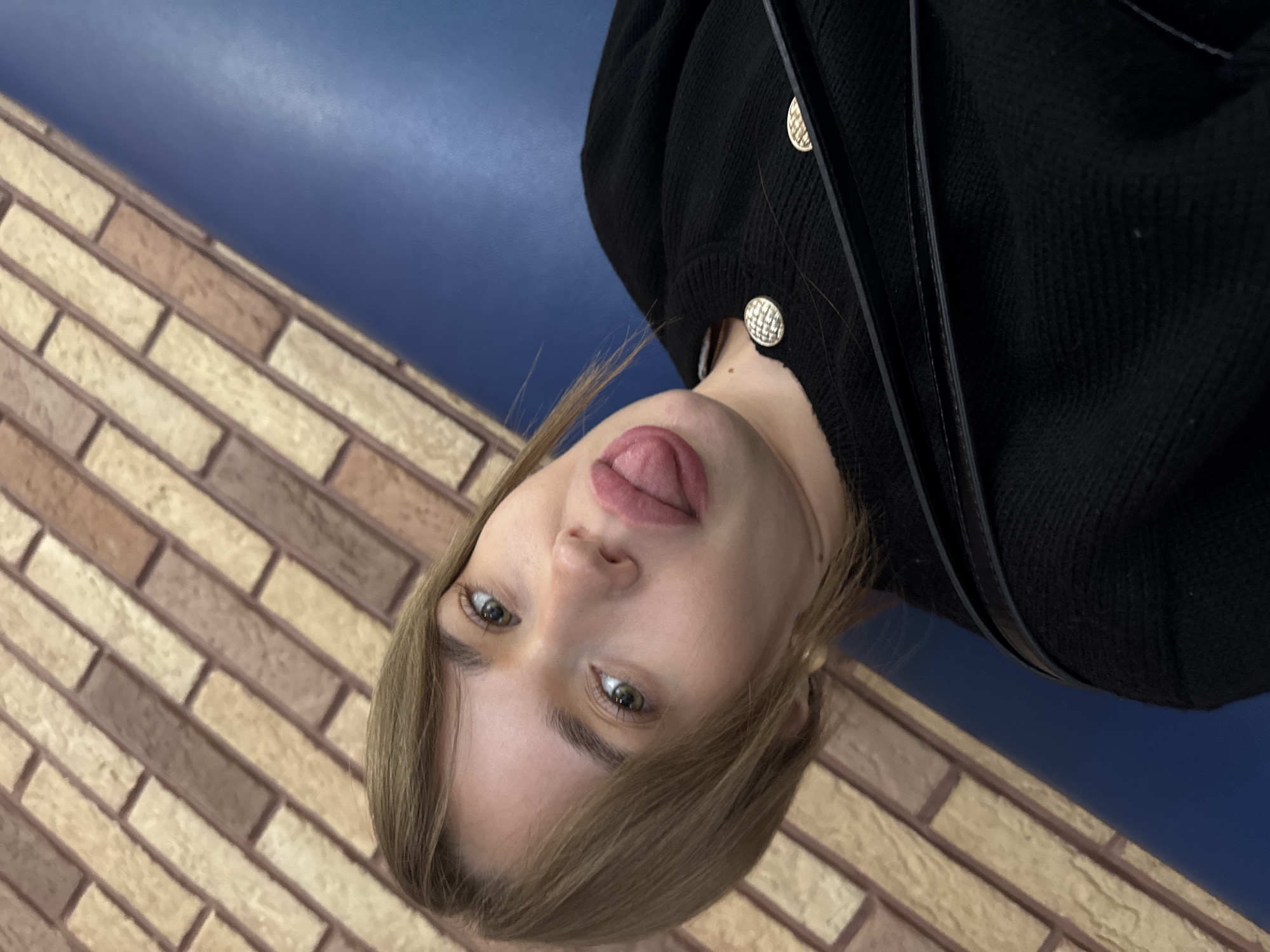 A woman with light brown hair and green eyes is sticking her tongue out towards the camera. She is wearing a black cardigan with gold buttons and a black purse strap is visible. The background consists of two sections: a brick wall with varying shades of brown and a smooth blue wall. The photograph appears to be taken indoors, and is rotated. The city of Chaikovsky, Russia, is referenced.