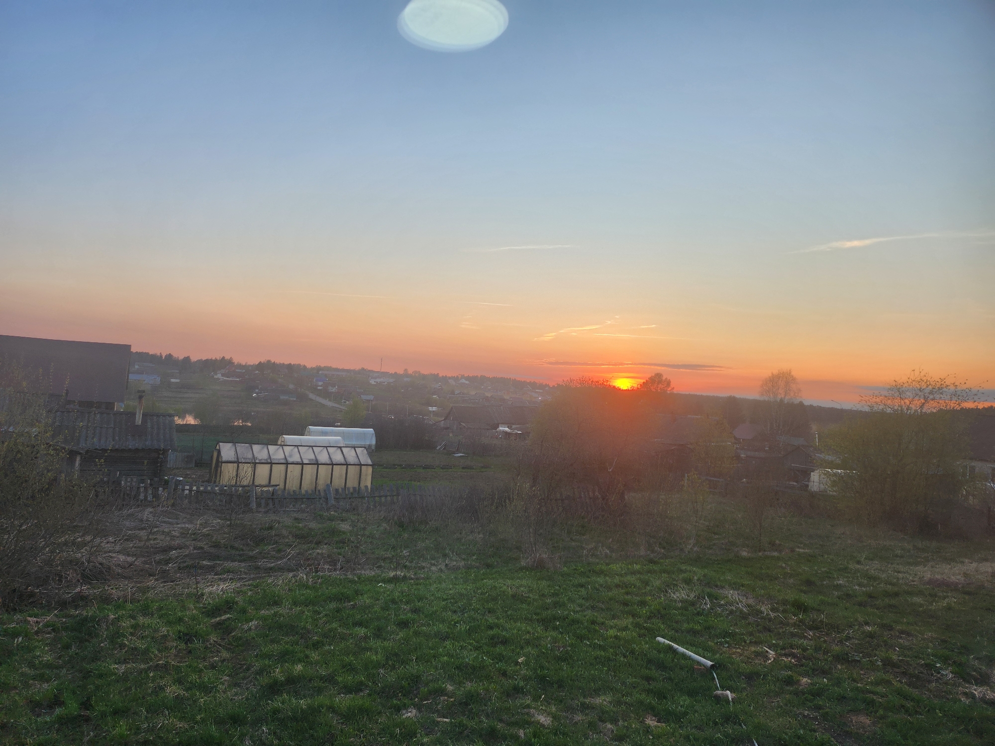 The image shows a rural landscape at sunset, likely in the vicinity of Krasnogorskoye, Russia, given the presence of structures consistent with Russian villages. In the foreground, there is a grassy field with varying shades of green. A discarded pipe is seen resting on the grass. In the midground, a fence partially obscures dilapidated wooden structures and multiple greenhouses. Beyond the midground, residential buildings make up the village. The horizon is lit by the setting sun, creating a gradient of orange, pink, and blue in the sky. A lens flare or reflection is present in the upper part of the image.
