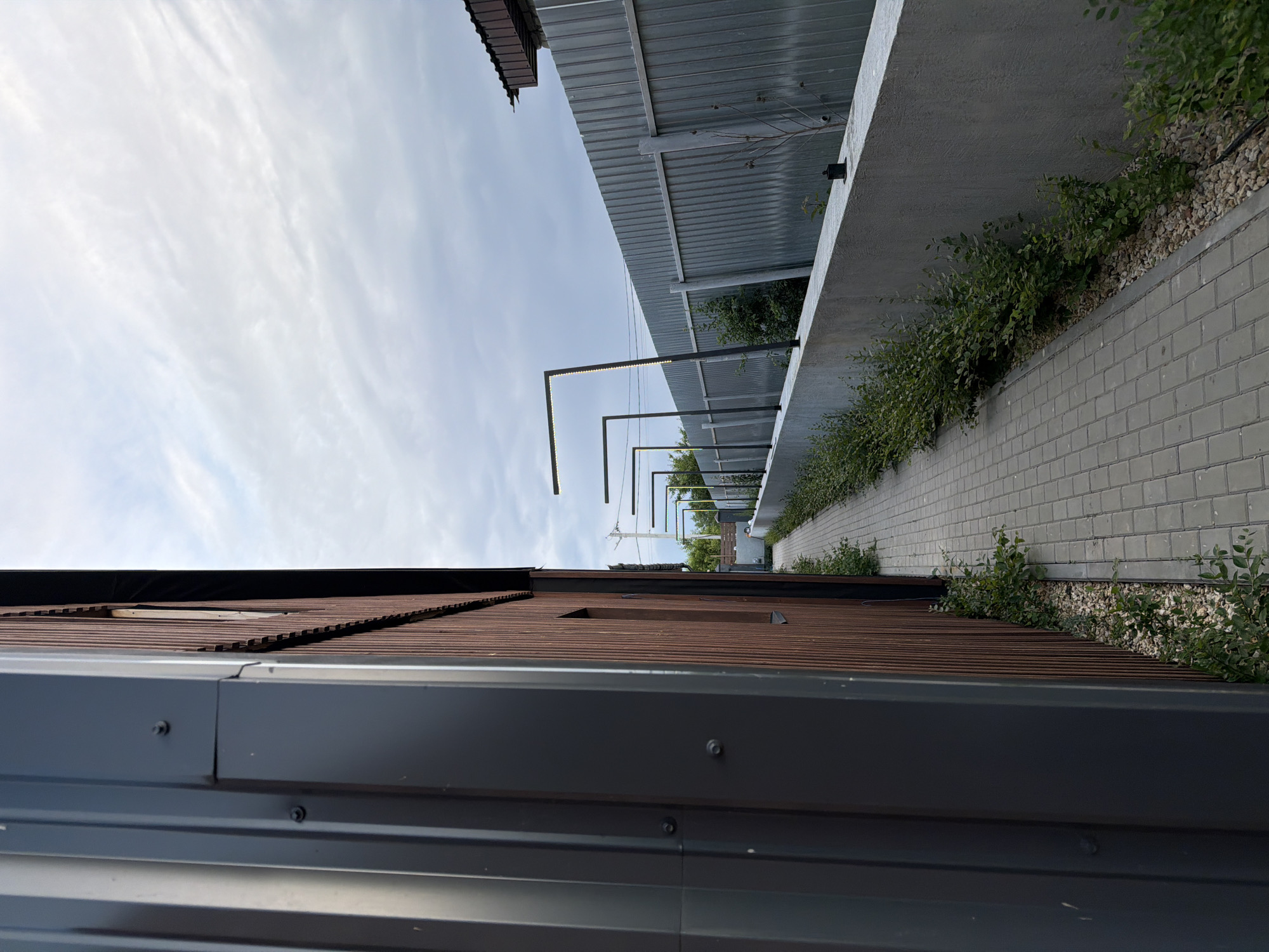 The image depicts a narrow paved walkway running alongside a building with vertical brown wooden siding and a dark grey corrugated metal downspout. The walkway is bordered on the right by a low concrete wall, beyond which is a tall corrugated metal fence. Several modern, angular light fixtures with LED strips are mounted on poles along the fence line. Green bushes and small trees grow between the concrete wall and the fence. The sky is overcast with scattered clouds. The scene appears to be an outdoor area, possibly a pathway or alleyway, in the city of Semiluki, Russia, given the context. There are no people visible in the image.
