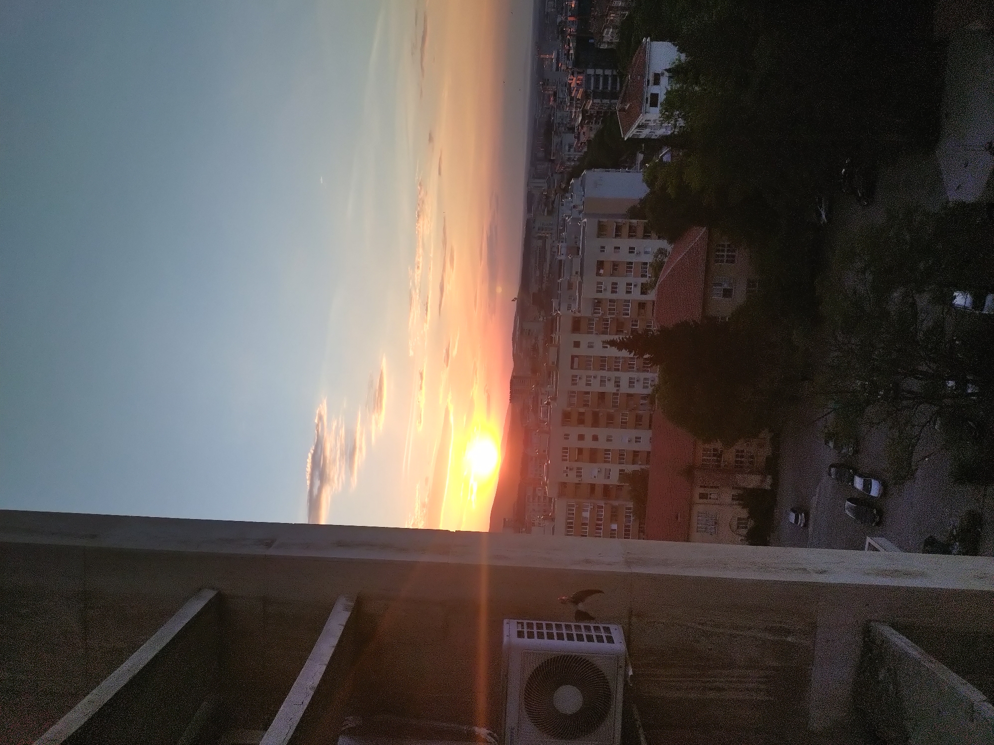 The image shows a sunset view from a rooftop in Stara Zagora, Bulgaria. In the foreground is a concrete rooftop structure, with an air conditioning unit visible on the right side. The background features a cityscape bathed in the orange glow of the setting sun. Buildings are densely packed in the central part of the view, transitioning to trees and vegetation on the right. The sky is a gradient of blue to orange, with a few clouds present. The overall perspective is looking down and out over the city from an elevated position.
