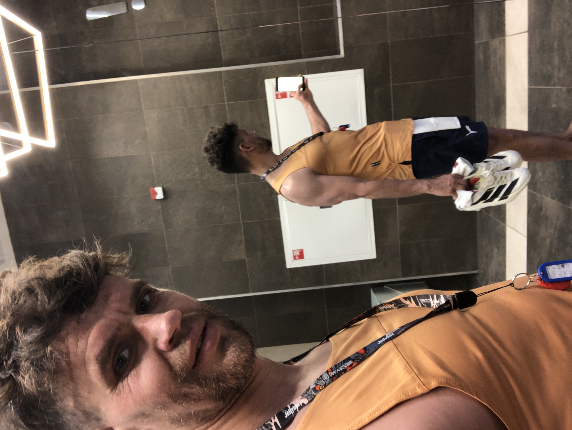 The foreground presents a close-up selfie of a male subject in what appears to be a modern changing facility in the city of Saint Petersburg, Russia. The man, approximately mid-30s to early 40s, possesses light brown, tousled hair with shaved sides and a beard. He wears an orange sleeveless tank top and a lanyard around his neck, featuring a multi-colored design and the text 'JellyBox,' from which blue and red key fobs are suspended. His muscular right arm is partially visible. The same individual is reflected in a large, vertical mirror directly behind him, offering a side profile view. In the reflection, he is dressed in the identical orange tank top and lanyard. He wears navy blue athletic shorts with a white vertical stripe on the left leg and a white Puma logo. He is barefoot, holding a pair of white Adidas athletic shoes with black stripes and white socks in his left hand. His right hand holds a smartphone with a brightly lit, blank white screen, pointed towards himself, consistent with taking a mirror selfie. The environment consists of dark grey, large-format tiled walls and a floor with a dark and light grey checkered pattern. A white rectangular panel with a small red upper-left indicator is mounted on the wall behind the reflected figure. Above this panel, a smaller red rectangular object is also affixed to the wall. The scene is illuminated by overhead geometric light fixtures.
