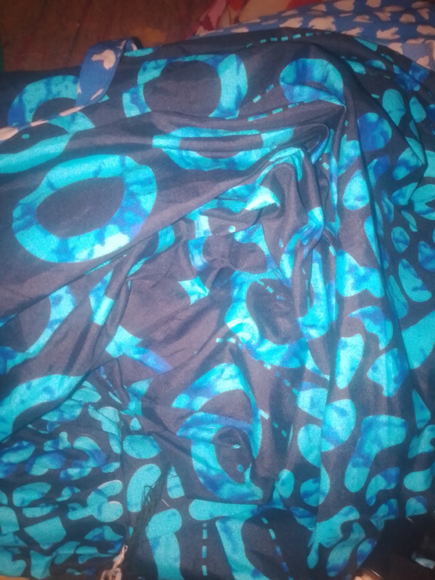 The image shows a close-up, overhead view of a dark blue fabric with a pattern of irregular, rounded shapes in various shades of turquoise and blue. The fabric is draped and folded, creating shadows and highlights that obscure some of the pattern. There are no people or identifiable objects other than the fabric itself. The background is blurry and out of focus. There is no information about the location of Jauro Yinu, Nigeria within the image or its context.