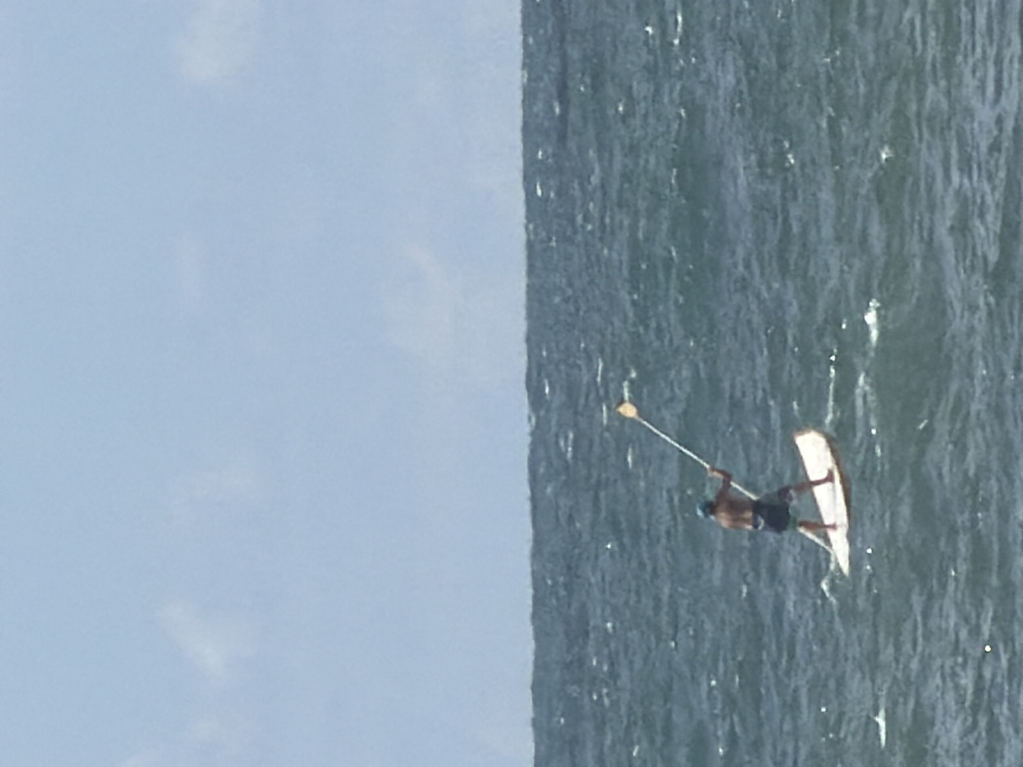 An outdoor scene depicts a lone individual stand-up paddleboarding on the Mediterranean Sea off Tel-Aviv, Israel. The upper portion of the image features a light blue sky with sparse, faint white cloud formations. Below the horizon, the water is a dark teal to grey-blue, characterized by numerous small waves and whitecaps, indicative of moderately choppy conditions.
A male individual, shirtless, wearing dark swim shorts with a possible patterned side, and a blue cap or bandana, stands centrally on a light-colored, elongated paddleboard. He holds a long, light-colored paddle with both hands, positioned over his right side, appearing to be in the motion of a stroke. The paddleboard is light-colored, possibly white or tan, and is actively riding a small wave. The scene is brightly illuminated, consistent with daytime.