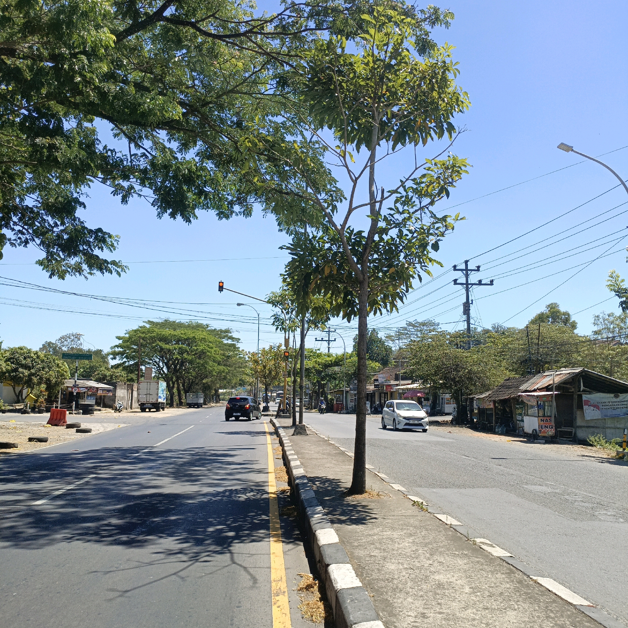The image depicts a street scene in Salatiga, Indonesia, on a bright, sunny day. The road is paved and divided by white lane markings. On the right side of the road, there is a sidewalk with a distinct black and white striped curb. Small trees line the sidewalk. Beyond the sidewalk, there are several small buildings with corrugated metal roofs, some appearing to be roadside stalls or shops. One stall has text that includes "NASENG T". A white car is traveling on the road, and further down, a black car is visible. A motorcycle is parked near one of the stalls. On the left side of the road, there are more trees, some with dense foliage, and a traffic light is visible in the distance. Power lines crisscross the sky, supported by utility poles. The sky is clear and blue. The overall impression is a moderately busy street in a tropical or subtropical urban environment.