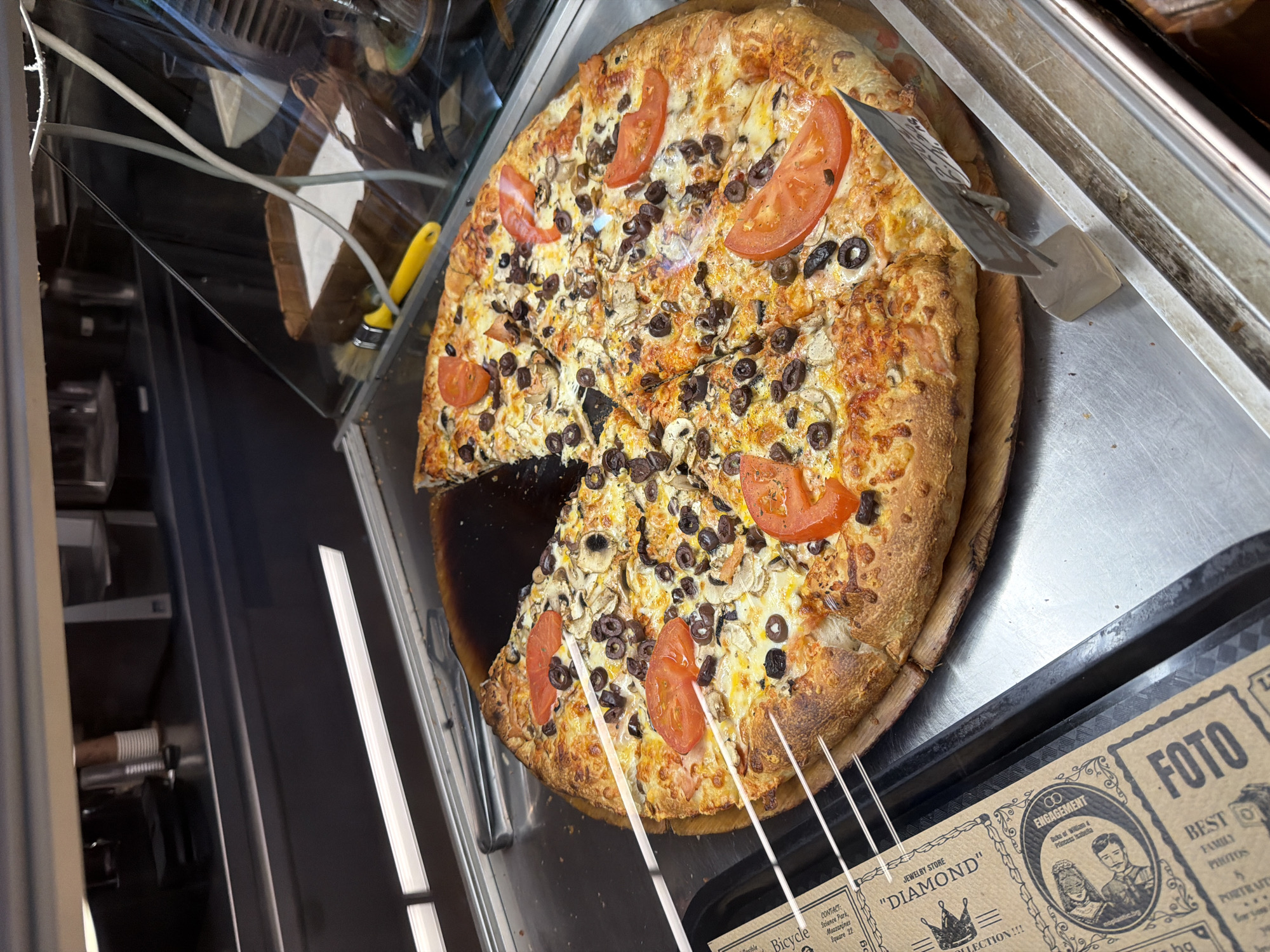 Here is a forensic description of the image:
The image depicts a pizza on a wooden board, with a slice removed. The pizza is topped with cheese, mushrooms, olives, and tomato slices. A metal spatula is present. The pizza sits inside a glass display case with a yellow-handled brush and wiring visible behind it.
In the lower right corner, portions of display cards are visible, with text including "FOTO", "BEST FAMILY PHOTOS", "ENGAGEMENT", "JEWELRY STORE", "DIAMOND", "Bicycle Science Park, Mezzanines Square 22".
The location is identified as Science Park, Mezzanines Square 22 in Unknown, Unknown.