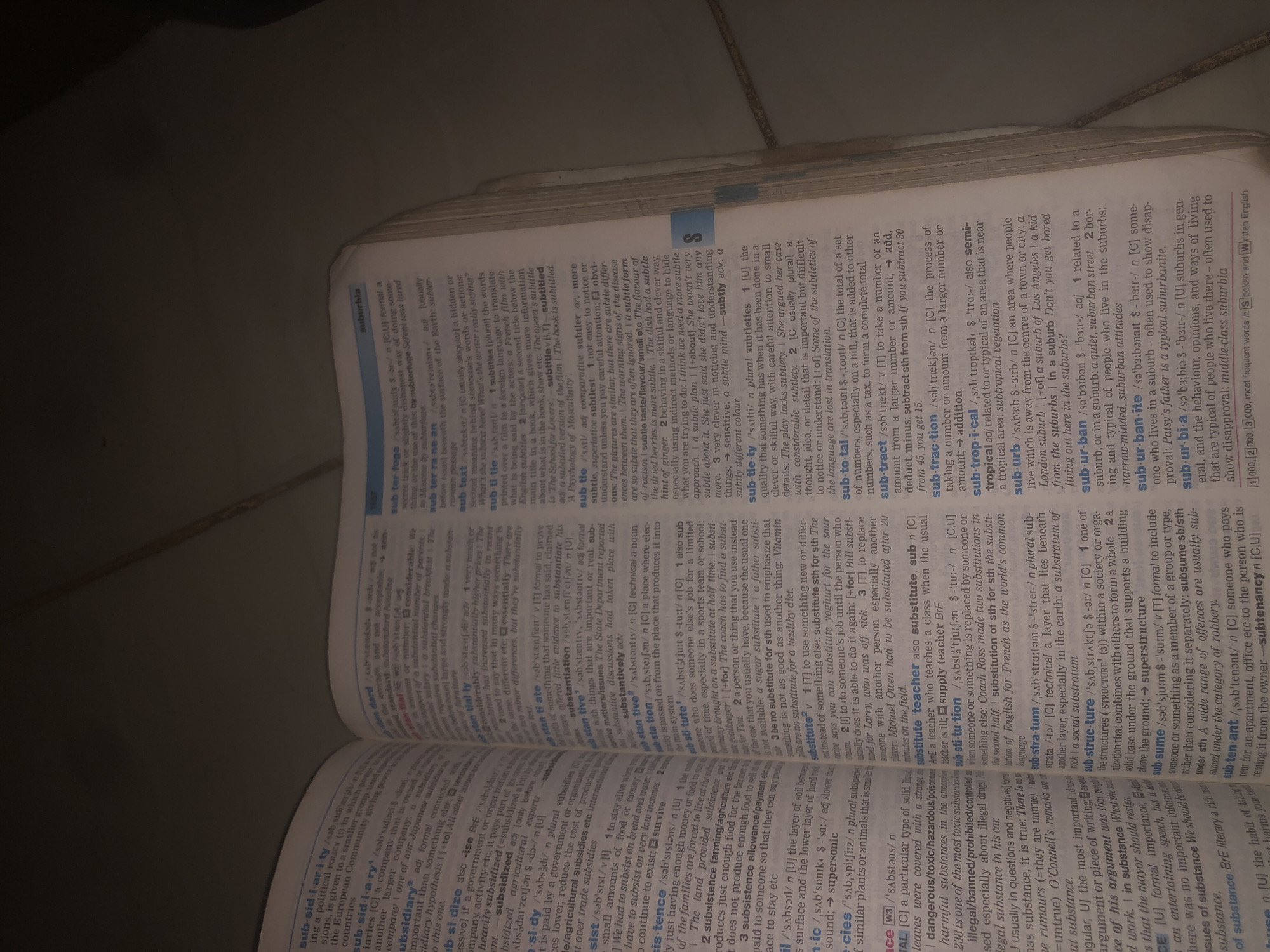 The image shows an open dictionary. The pages are filled with text, defining various words. The text is small and densely packed, with some words highlighted in bold. The dictionary appears to be well-used, with some pages slightly dog-eared. The lighting in the image is dim, suggesting it might be nighttime or a poorly lit room. There is no indication of the city of Jalingo, Nigeria, or any of its landmarks in the image. The context is limited to the interior of a room where a person is reading or consulting a dictionary.