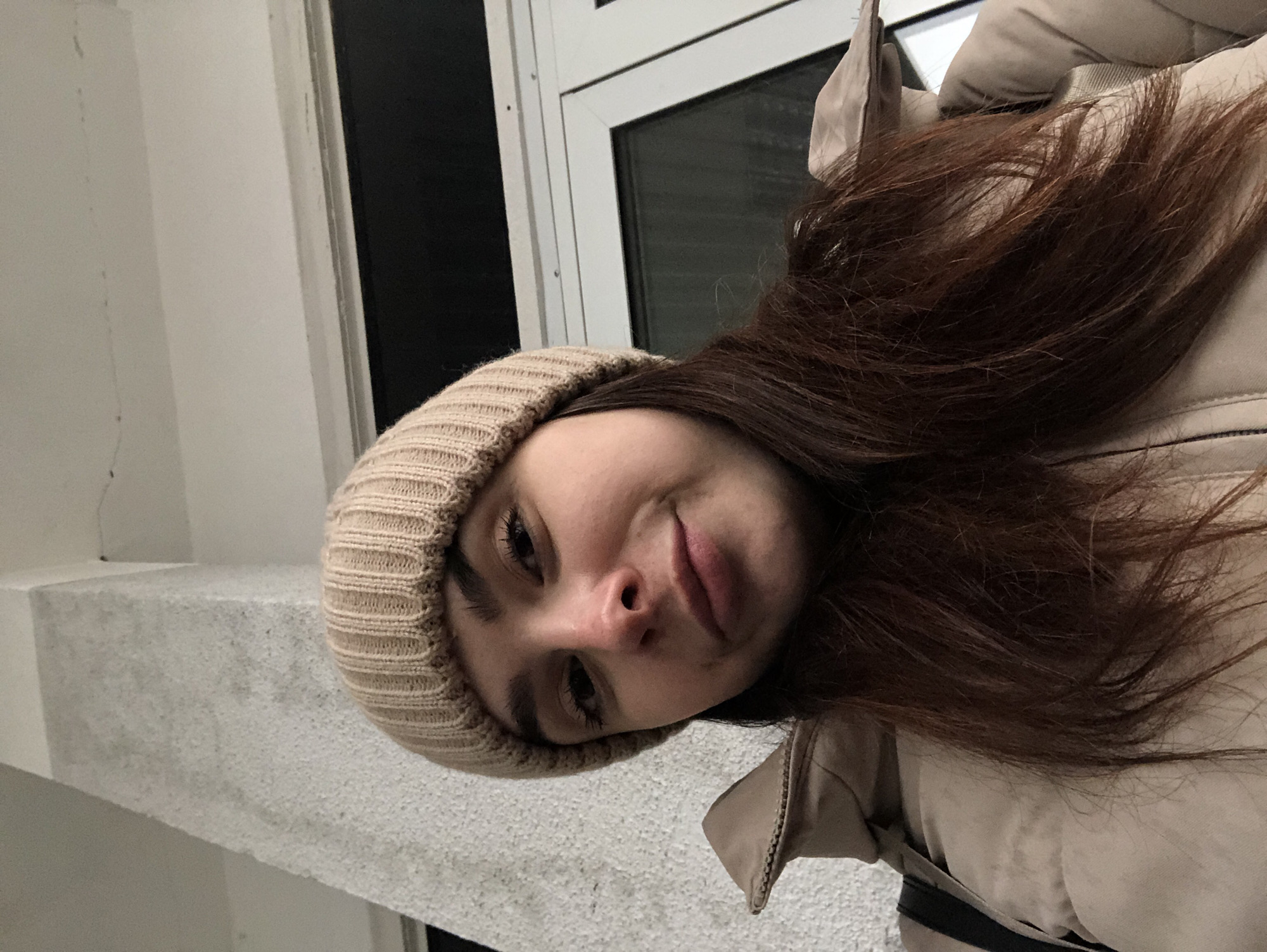 A young woman wearing a beige knit beanie and a beige jacket poses for a selfie. She is indoors, near a white ledge and a window with a black frame. The location appears to be in Russia, possibly the city of Chaikovsky.