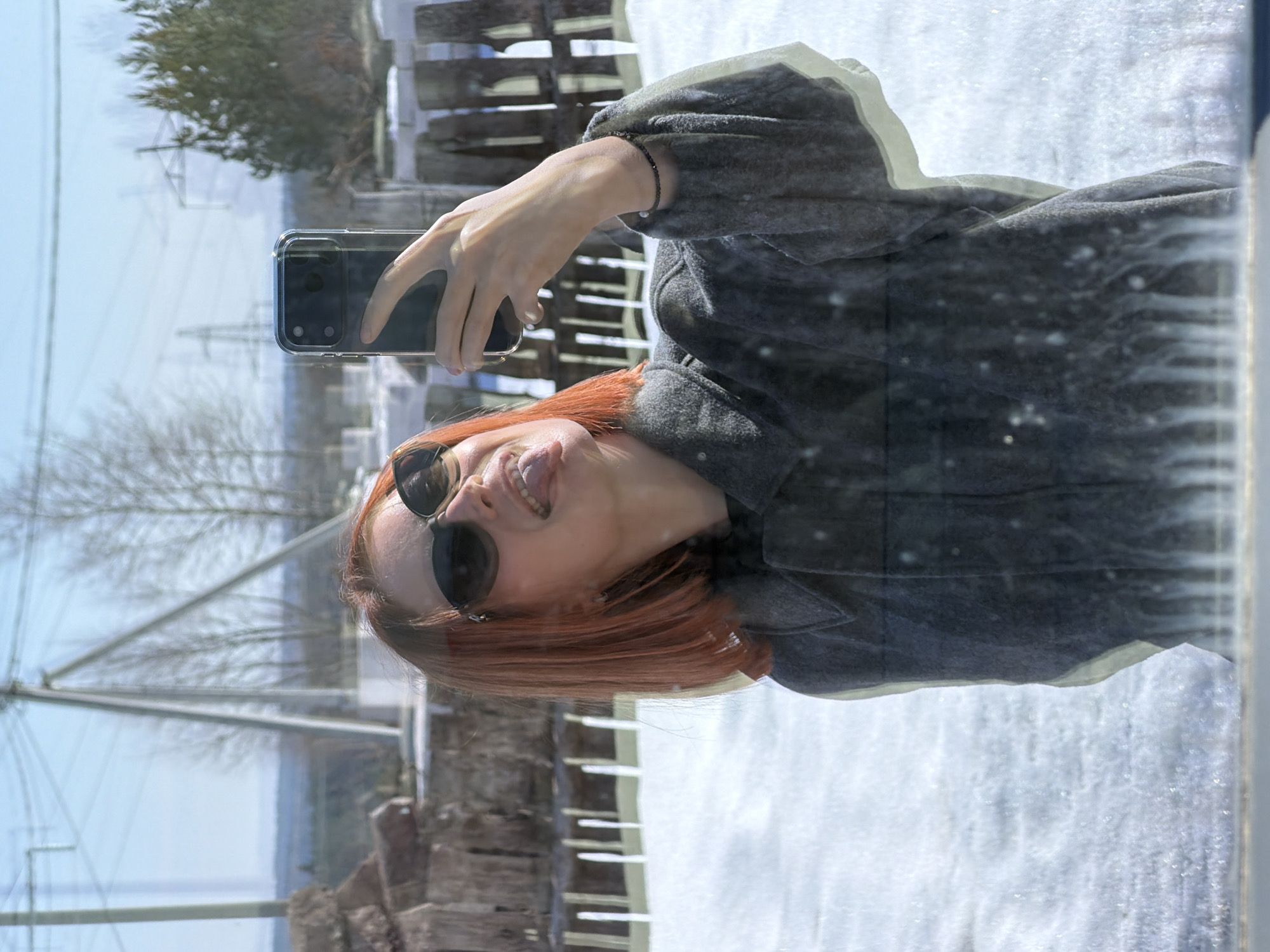 A woman is taking a selfie in front of a window in Chaikovsky, Russia. She is wearing sunglasses and sticking out her tongue. The scene visible through the window includes trees, a wooden fence, snow-covered ground, power lines, and distant buildings. The photograph is taken with a phone in a transparent case. The image is a reflection of the woman and the surrounding environment in the glass. The location has snow on the ground and appears to be outdoors.