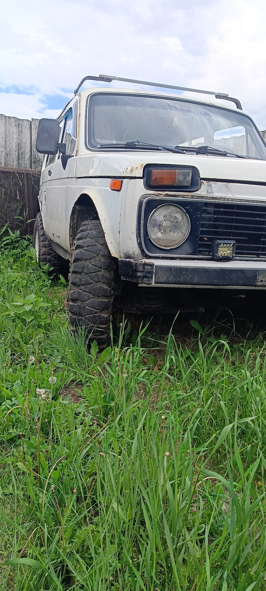 An off-road vehicle, identified as a white Lada Niva, is parked in a grassy area next to a weathered grey fence. The vehicle appears to be in a state of disrepair, with visible rust on its exterior and dirty tires. The front of the vehicle is angled towards the viewer, showcasing its large, rugged tires and a aftermarket light bar mounted on the front bumper. The surrounding environment is overgrown with tall green grass and some weeds, suggesting a rural or undeveloped location. The sky is partly cloudy. This scene is located in the village of Kamennoye, Russia.
