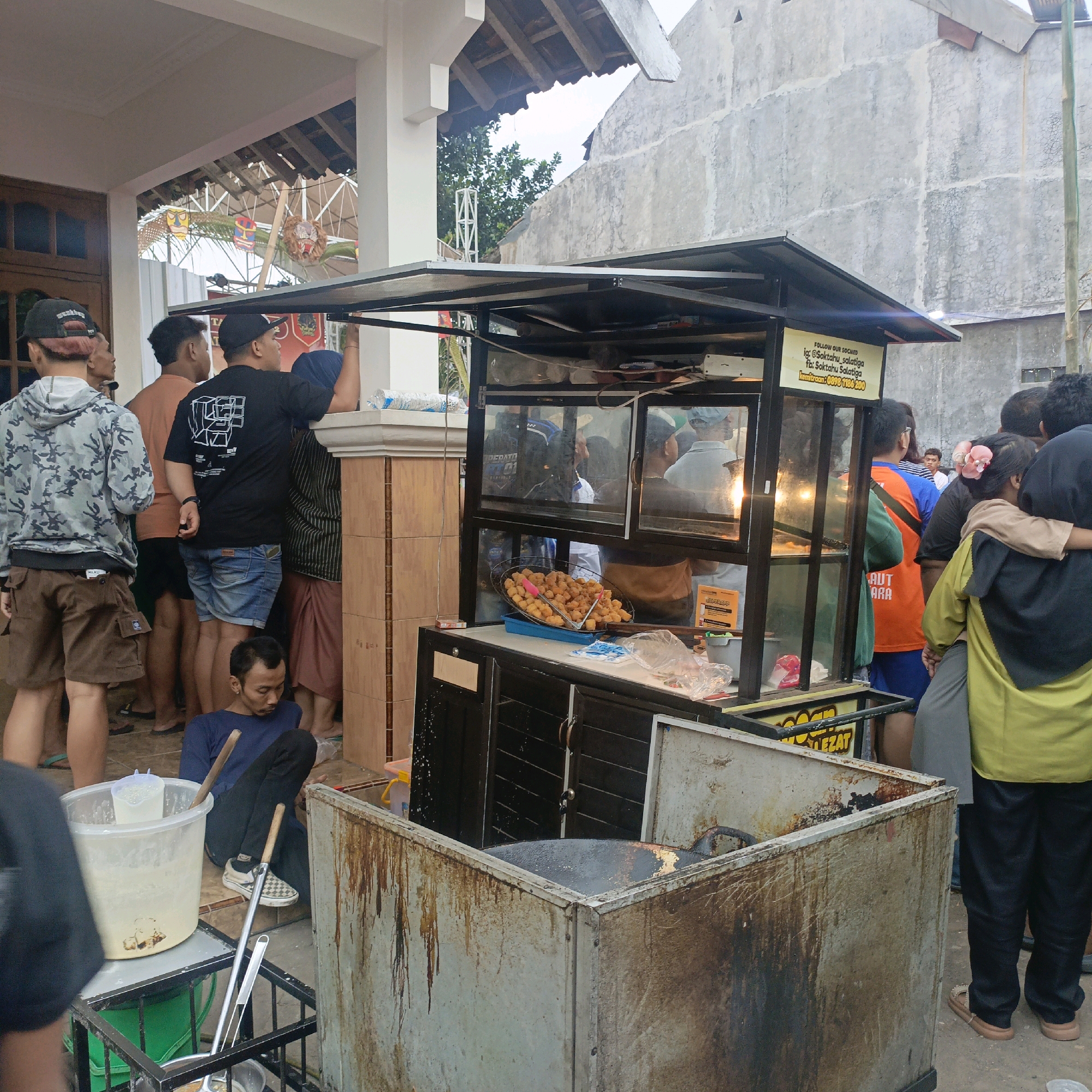 The image captures a street food stall in Salatiga, Indonesia. A food cart, made of metal and glass, is the central focus. The cart is filled with food, likely snacks, displayed in a wire basket. Behind the cart, a group of people are gathered, some appearing to be customers and others involved in the food preparation. One individual, wearing a blue shirt and black pants, is seated on the ground next to the cart. The background shows a building with a textured concrete wall, a rooftop, and some greenery. A sign on the cart includes social media handles, "Soktahu_Salatiga," and a contact number, indicating the stall's location or affiliation with Salatiga. The scene appears to be a busy street food operation, with people interacting and food being served.