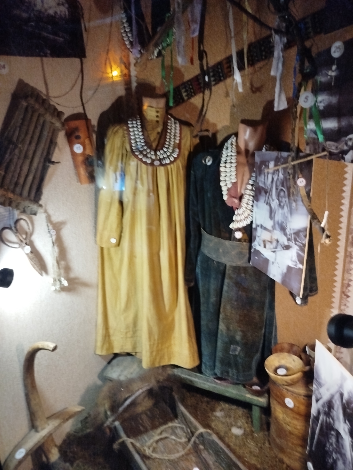 The image displays an exhibit, likely within a museum, showcasing cultural artifacts. In the foreground, a wooden tool with a curved handle rests on what appears to be straw or dry grass. Behind it, a long, narrow wooden vessel with rope is visible, also on the straw.
Dominating the center of the image are two mannequins dressed in traditional clothing. The mannequin on the left wears a long, yellow garment adorned with a shell necklace. To its right, another mannequin is dressed in a dark garment, also featuring a shell necklace, and a dark belt cinches its waist. A large, framed photograph is suspended in front of this mannequin.
The background consists of a textured wall, decorated with various cultural items. These include woven materials, hanging fabrics, and what appear to be decorative elements made from shells or stones. Several numbered circular markers are affixed to the wall and some of the objects. A small lamp with a warm yellow light is positioned on the wall between the woven material and the yellow garment.
This exhibit is located in the city of Tashtagol, Russia, as indicated by the contextual information provided.