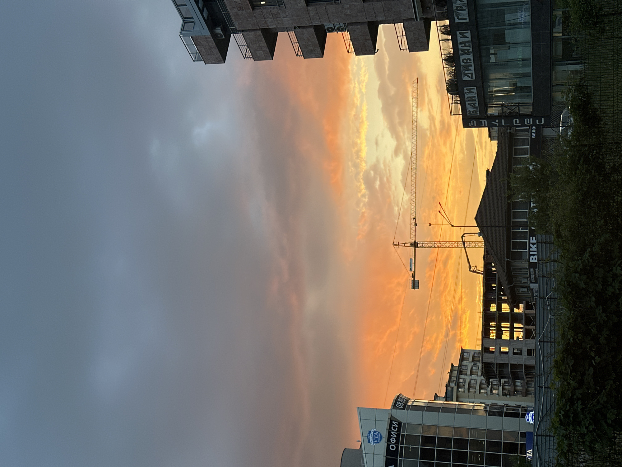 The image depicts an urban landscape in Sofia, Bulgaria, taken during a vivid sunset. The sky is a gradient of colors, transitioning from a stormy gray on the left to vibrant orange and yellow near the center. A construction crane stands prominently in the middle of the frame, silhouetted against the colorful sky. Various buildings are visible, ranging from modern glass-fronted structures to older buildings with signs in Cyrillic script. Some of these signs include the word "ОФИСИ" (offices), "БАНКИ" (banks), "ДИВ ИН" (DII IN) and "БАЙК" (BIKE) (bike). Green trees and bushes partially obscure some of the buildings on the right side.