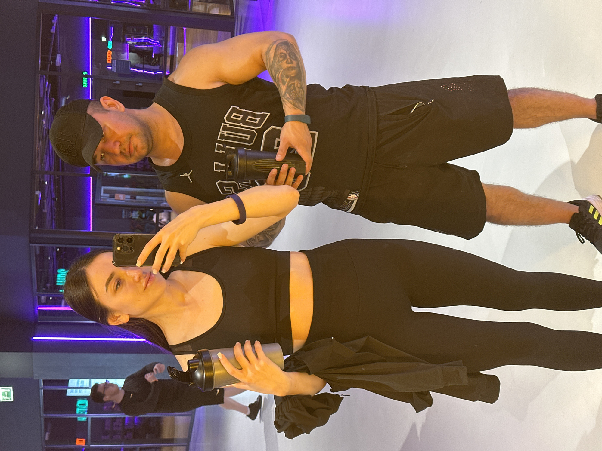 The image captures a couple in a gym, likely in Kazan, Russia, given the visible Cyrillic signage in the background. The woman, on the left, is taking a selfie with her phone. She is wearing a black sports bra and black leggings, holding a shaker bottle in her left hand. A black jacket is draped over her left arm. The man, on the right, is wearing a black tank top with white lettering and a black baseball cap. He is also holding a shaker bottle. Both individuals are wearing fitness attire, suggesting they are either working out or have just finished. The gym environment is suggested by the mirrored walls and equipment visible in the background, with purple lighting adding to the atmosphere. Another person, a man wearing glasses and a black long-sleeved shirt, is visible in the background, slightly out of focus.