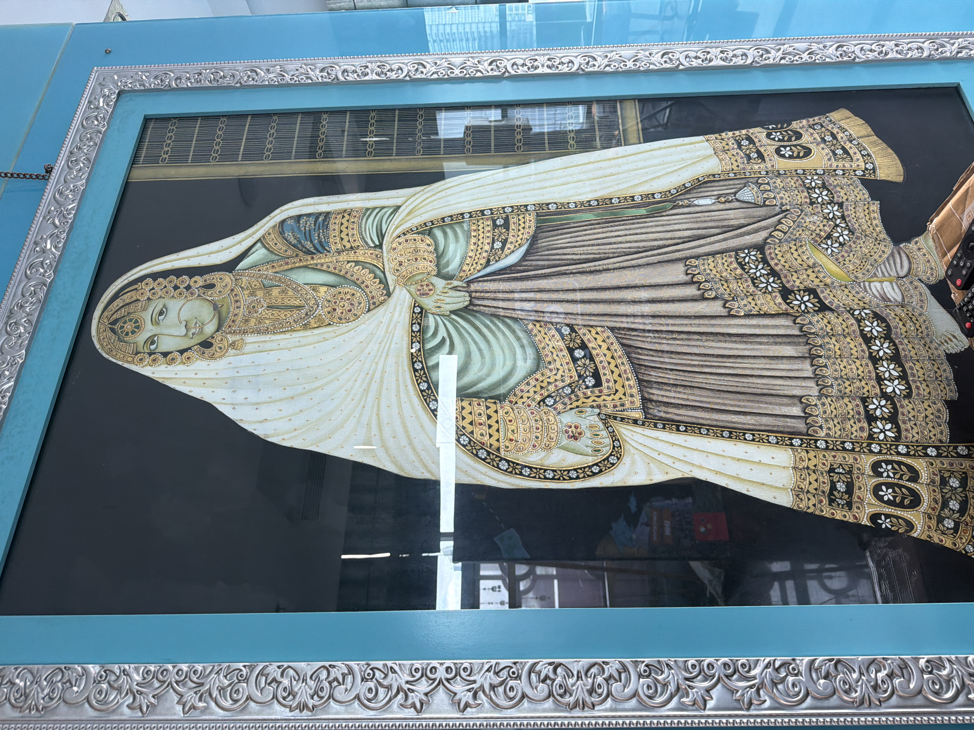A framed portrait of a woman is displayed inside a building in South Jakarta, Indonesia. The portrait features a woman in traditional attire, including a white head covering and ornate jewelry, set against a dark background. The frame is two-toned, with a light blue exterior and a decorative silver interior trim. Reflections on the glass surface of the frame indicate an indoor setting with visible elements of the building's interior.