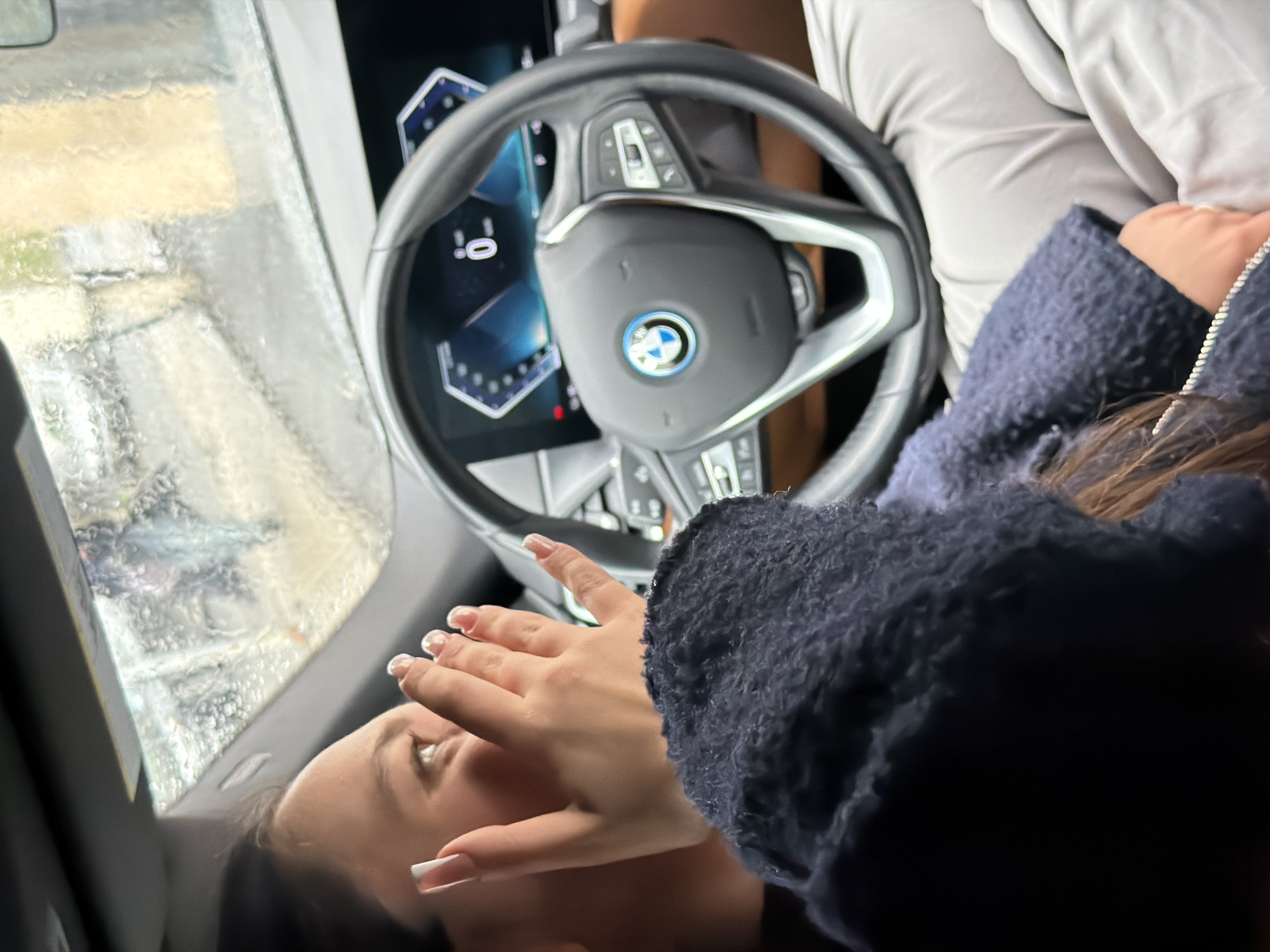 The image depicts the interior of a modern BMW vehicle, likely in motion or stationary during rain. The viewpoint is from a passenger position, looking forward through the front windshield, which is heavily streaked with raindrops. A female individual occupies the left foreground, with her right profile visible, including her right eye and cheek. She has dark hair and her right hand, adorned with long nails featuring white tips, is raised towards her face, partially covering her mouth. She wears a dark blue, textured, hooded garment. Her gaze is directed out the windshield to the right. The vehicle's black and silver steering wheel, centered with a prominent BMW logo, is in the midground. Behind the steering wheel, a modern digital instrument cluster displays a "0" (likely indicating speed) along with other blue graphical elements. Brown interior accents are visible beyond the steering wheel. Through the rain-covered windshield, a blurred urban environment is discernible, including indistinct architectural forms, with one building appearing light-colored. The specific location within the city of Yerevan, Armenia, cannot be determined from the visual information provided.