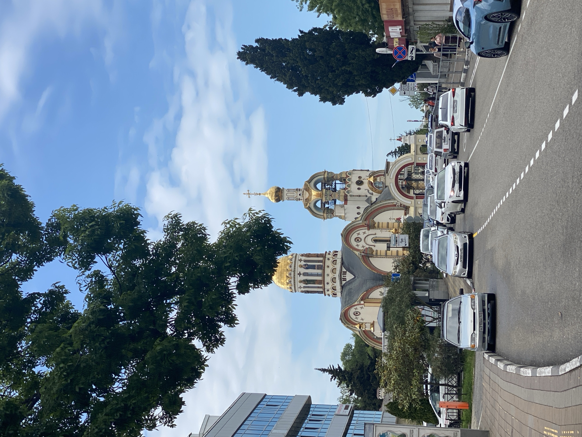 The image shows a street scene in Sochi, Russia, featuring the Cathedral of Archangel Michael. A line of parked cars is present on the side of the street closest to the cathedral. A sidewalk and various trees are visible along the edge of the road. A modern, multi-story building is partially visible at the bottom left. The sky is partly cloudy. Some people can be seen on the pedestrian crosswalk.