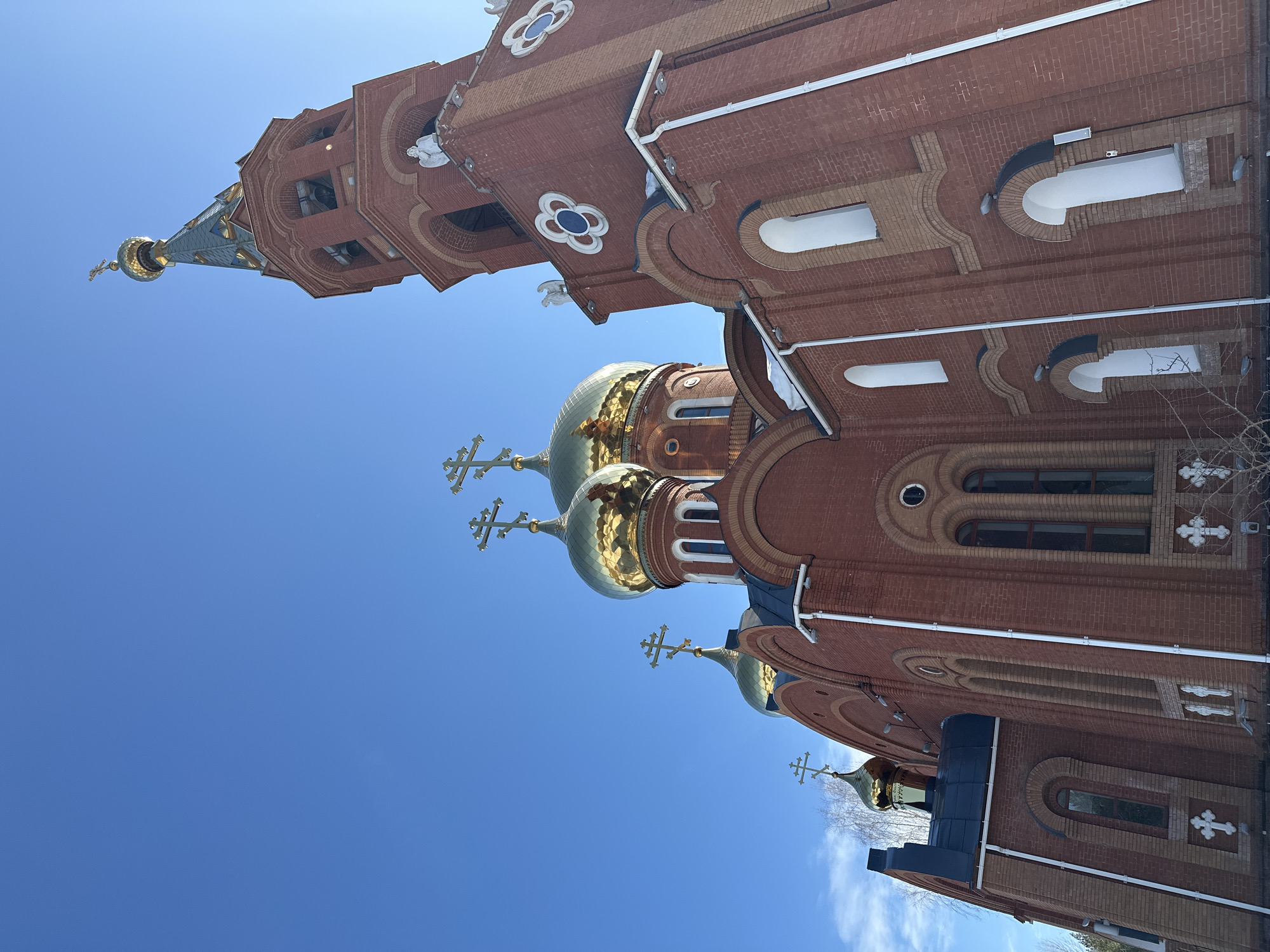 This image displays an Orthodox church located in Chaikovsky, Russia, under a clear, bright blue sky. The primary structure is constructed of red brick, featuring several golden onion domes, each topped with a golden Orthodox cross. A prominent, multi-tiered red brick bell tower stands to the right, crowned with a dark-colored, scale-patterned spire that culminates in a smaller golden onion dome and cross. The bell tower incorporates arched openings revealing bells and is adorned with small white angel figures positioned on its upper corners. The church's facade includes multiple arched windows, some recessed with white interiors, and decorative white clover-shaped medallions embedded in the brickwork. White rain gutters and downspouts are present along the building's exterior. Patches of snow are visible on some roof ledges. Bare tree branches are discernible in the lower-left background. No individuals, specific actions, or interactions are visible.