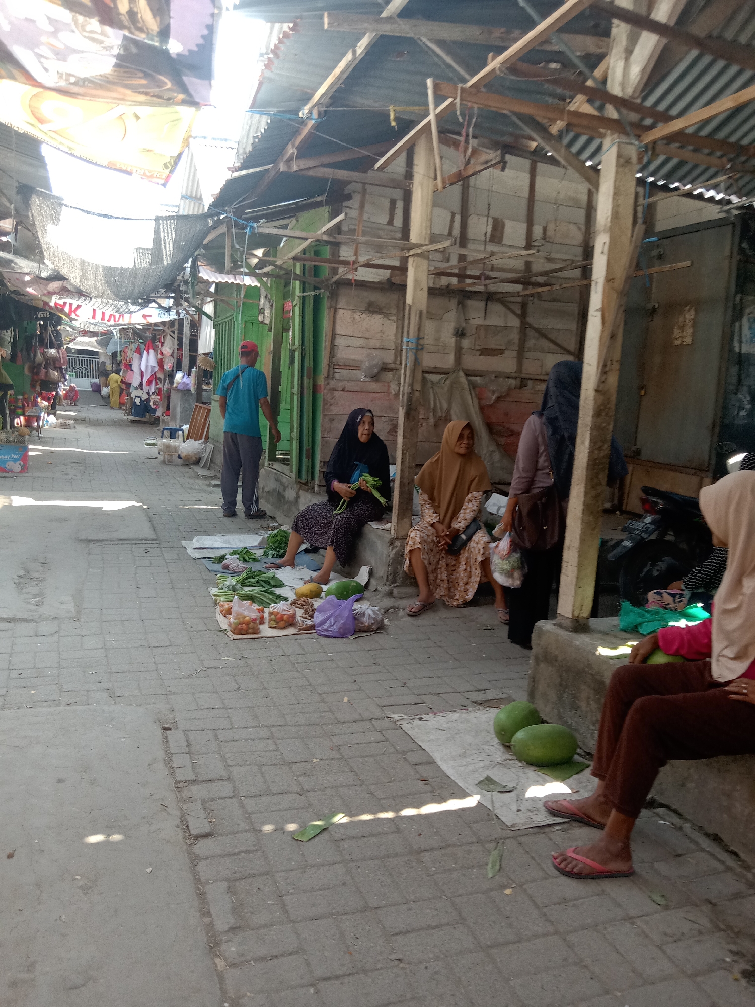 The image depicts an outdoor market scene in Klepek, Indonesia. In the foreground, a person wearing brown trousers and pink flip-flops is seated on a concrete ledge, with two large green gourds on a mat beside them. Further into the market, two women in hijabs are sitting on the ground near a stall. One woman, wearing a black hijab and dress, holds a bunch of green vegetables. The other woman, in a tan hijab and patterned dress, has her hands in her lap. A man in a blue shirt and gray pants stands near a green-walled structure, facing away from the camera. A variety of produce, including tomatoes and other vegetables, are displayed on mats on the ground. The market stalls are constructed with wooden frames and corrugated metal roofing, suggesting a semi-permanent setup. The ground is paved with interlocking bricks and concrete. In the background, there are more stalls and structures, with awnings and banners visible, indicating a bustling marketplace. The lighting suggests it is daytime, with bright sunlight casting shadows.