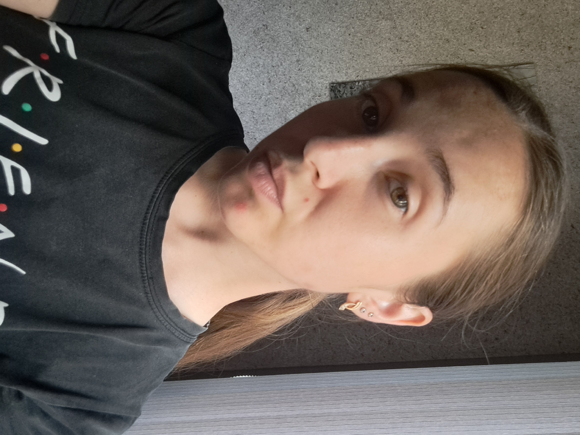 The image is a selfie of a young woman with light brown hair tied in a ponytail. She is wearing a black t-shirt with the word "FRIENDS" in white lettering, and is facing the camera, looking slightly to the side. She has two small stud earrings and one hoop earring in her left ear. There are two small red blemishes on her face, one on her cheek and one on her chin. The background is a textured wall and a vertical blinds. The image was taken in Vypasne, Ukraine.