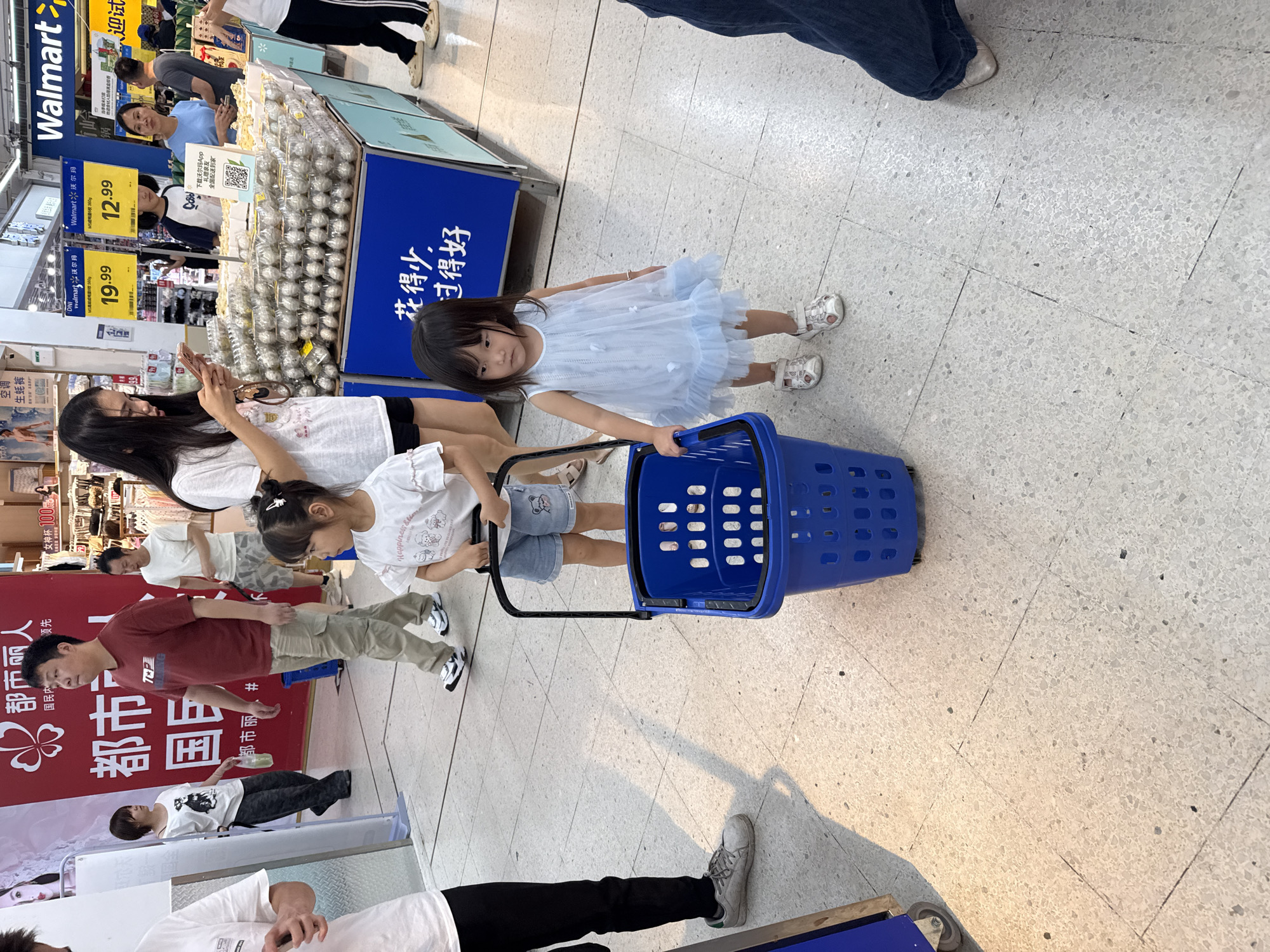 The image depicts an indoor scene, likely a supermarket, possibly in Huizhou, China, indicated by signage in Chinese. In the foreground, a young girl in a light blue dress and white sandals stands beside a blue shopping basket. To her left, another young girl in a white t-shirt and denim shorts holds the handle of the basket. Behind them, a woman in a white t-shirt is looking at her phone. Further back, a man in a maroon t-shirt and cargo pants walks past. Other shoppers are visible in the background, with shelves of products lining the store. The flooring appears to be tiled.