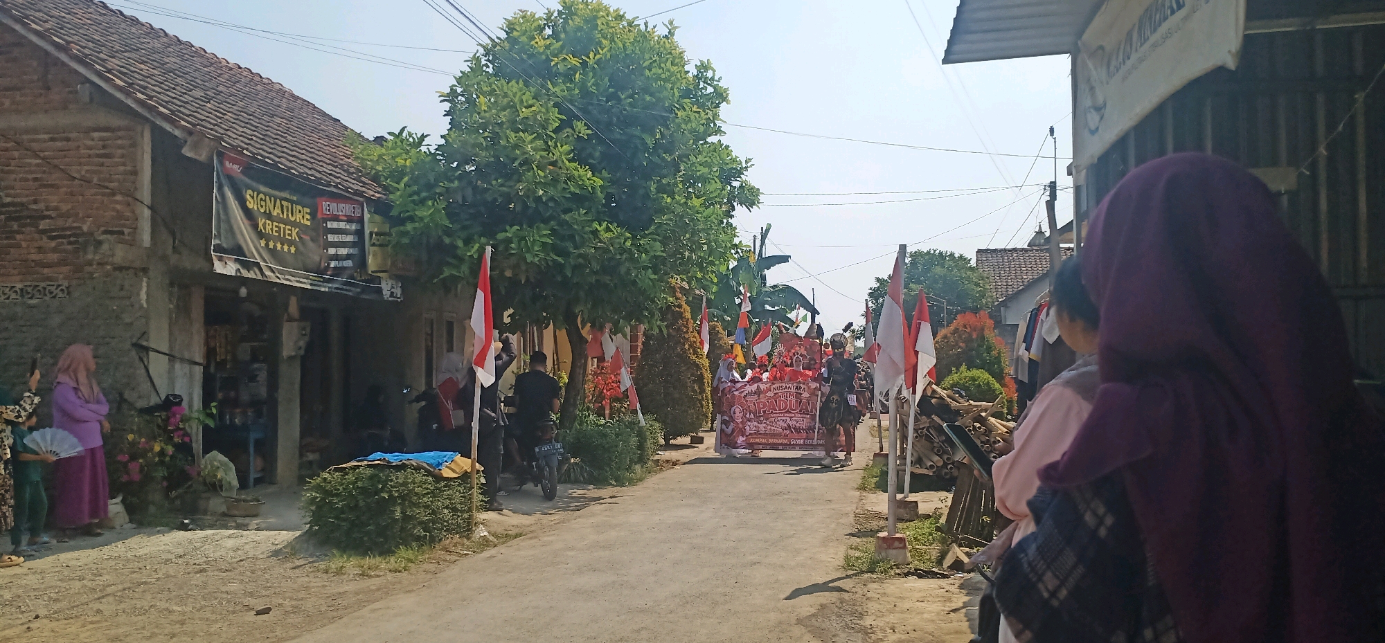 The image depicts a street scene in Kincang, Indonesia, likely during a procession or celebration. The road is paved and appears to be in a village or suburban area, with houses and shops lining either side. A group of people are walking down the street, carrying a large banner that reads "NUSANTARA ILMI PADUAN" and other Indonesian text. They are accompanied by several Indonesian flags. Some individuals are dressed in traditional attire, and one person is wearing a headdress. On the left side of the road, a few people are standing near a shop that has a sign with "SIGNATURE KRETEK" and stars. There are also motorcycles parked on the side of the road. Lush green trees and foliage are visible throughout the scene, and the sky is clear and bright.