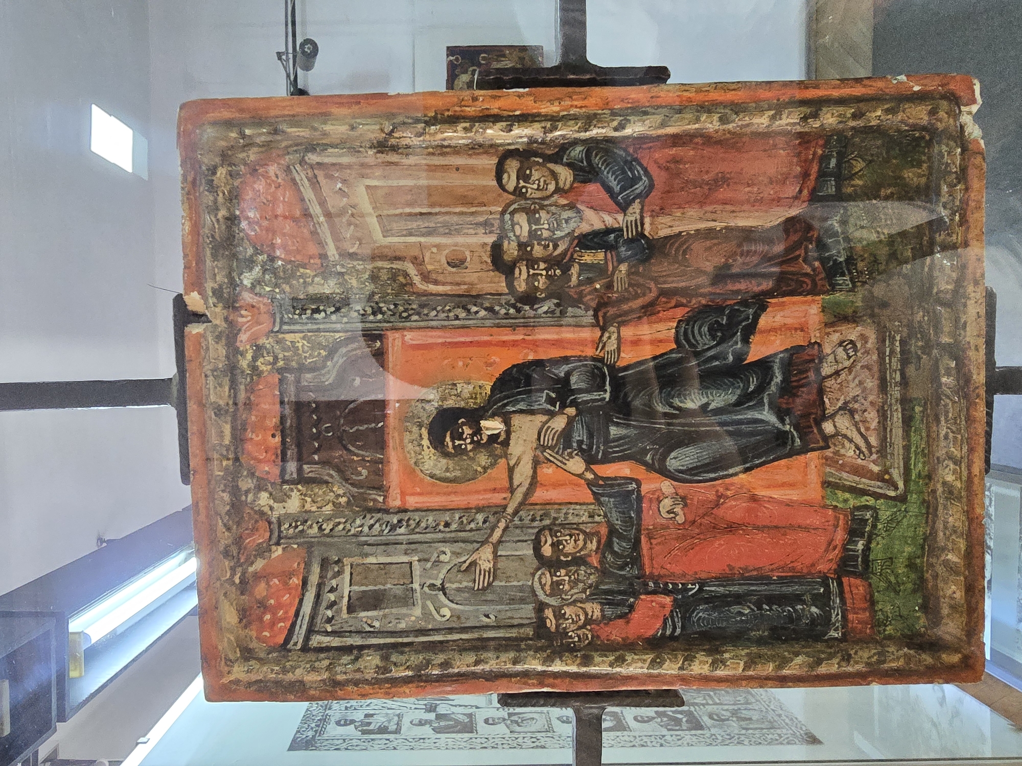A rectangular wooden icon panel, approximately 80-90 cm in height and 50-60 cm in width, is depicted behind a reflective glass surface within a museum setting. The icon, exhibiting significant craquelure, pigment loss, and visible cracks, primarily features a central religious scene with a warm, earthy color palette of ochre, sienna, deep red, black, and green.
The central figure, presumed to be Jesus Christ, stands barefoot on a green ground, clad in a dark blue or black robe with a reddish undertunic. A golden halo encircles his head. His right arm is extended outwards and slightly upward, palm open, while his left hand is positioned over his chest, with two fingers extended. He has a dark beard and hair, with prominent dark eyes.
To the left of the central figure, a group of five men is depicted. The foremost man, wearing a red tunic and dark robe, extends his left hand to touch the central figure's right forearm. Other figures in this group are similarly attired in tunics of red, brown, and black. Their faces are rendered with dark eyes and somber expressions, some with beards.
To the right of the central figure, a group of four men stands. They are dressed in tunics and robes of red and dark colors. The figure closest to the central figure appears to have a beard. Their faces also feature dark eyes and solemn expressions.
The background is defined by architectural elements, including two prominent doorways or arches in shades of gray and brown on the sides, and a central, more ornate archway above the central figure's head, set against a bright orange wall. The floor beneath the figures is a solid green.
The image is photographed through protective glass, which displays prominent reflections of overhead fluorescent lighting, particularly in the upper left and upper right portions of the frame. Reflections also show a ceiling, a white wall, and part of a metal display stand supporting the icon. To the far left, behind the icon, parts of framed monochrome photographs or prints are visible on an adjacent wall. This artifact is located in a museum in Teteven, Bulgaria.