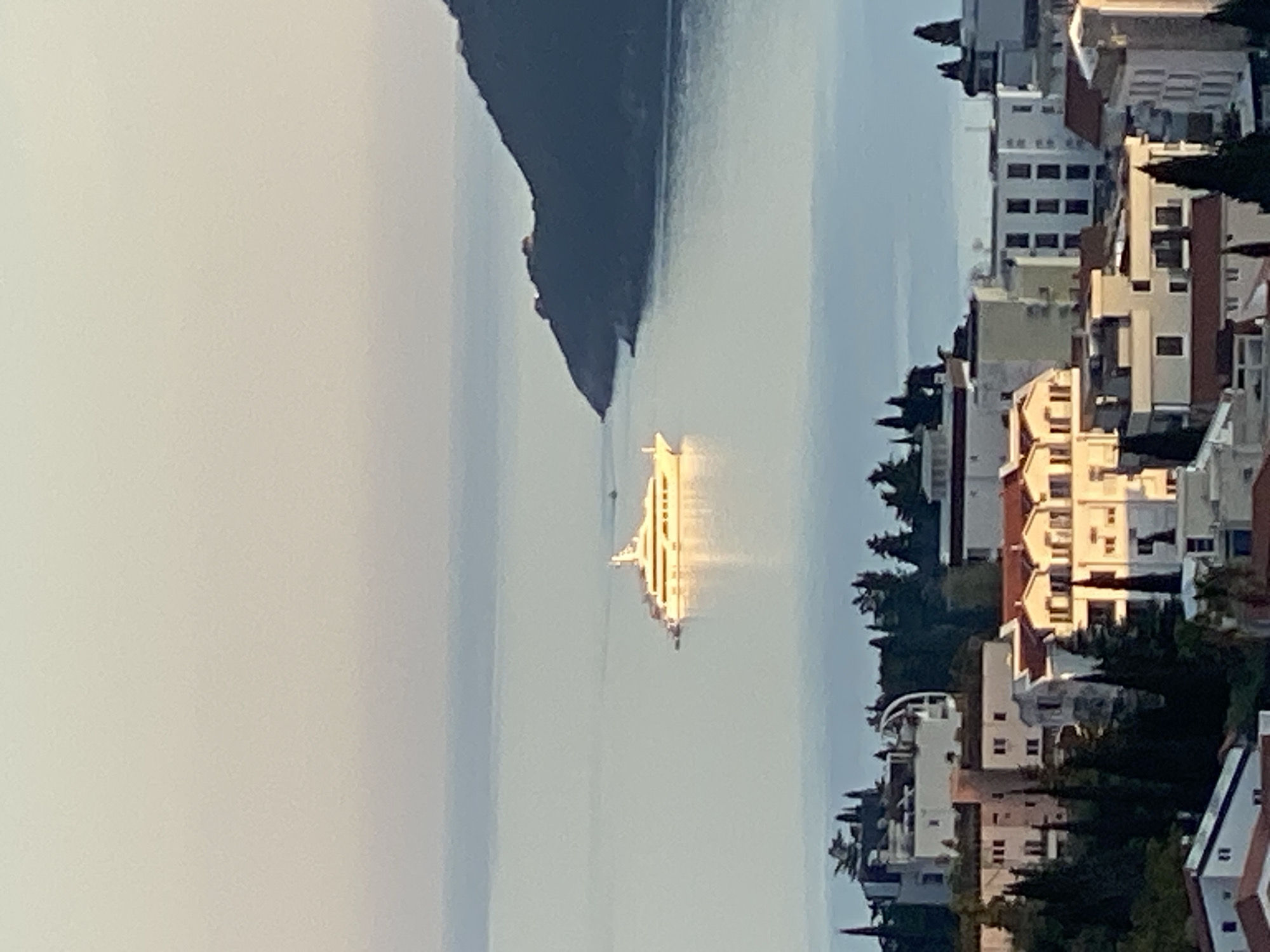 The image depicts a seaside landscape in Budva, Montenegro, featuring a large cruise ship on the water. Buildings with varied architectural styles and colors are arranged along the coastline, interspersed with trees. A dark mountainous landmass is visible on the left side of the frame. The scene is bathed in sunlight, which creates reflections on the water and brightens the ship. The sky appears hazy.