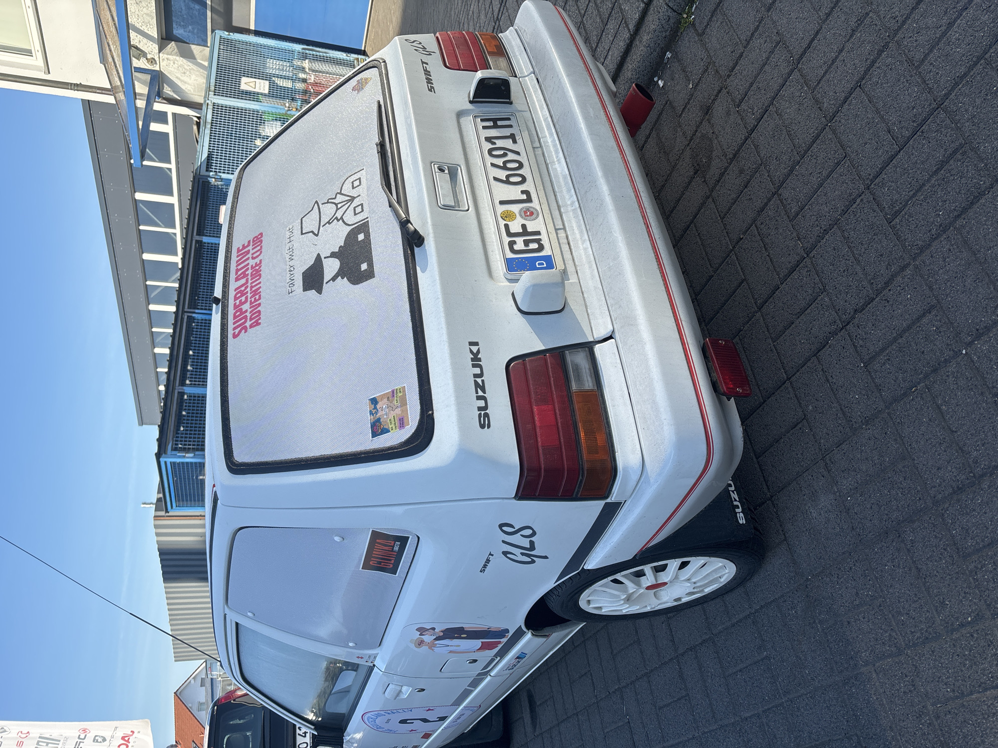 A white Suzuki Swift GLS hatchback is parked on a dark grey brick-paved road. The vehicle has a German license plate "GF•L 6691H." There are several stickers on the car, including one on the rear window that reads "SUPERLATIVE ADVENTURE CLUB" and "Fahrer mit Hut" (Driver with a Hat). The car has white wheels with red accents. The city of Fulda, Germany, is not explicitly identifiable in the image.