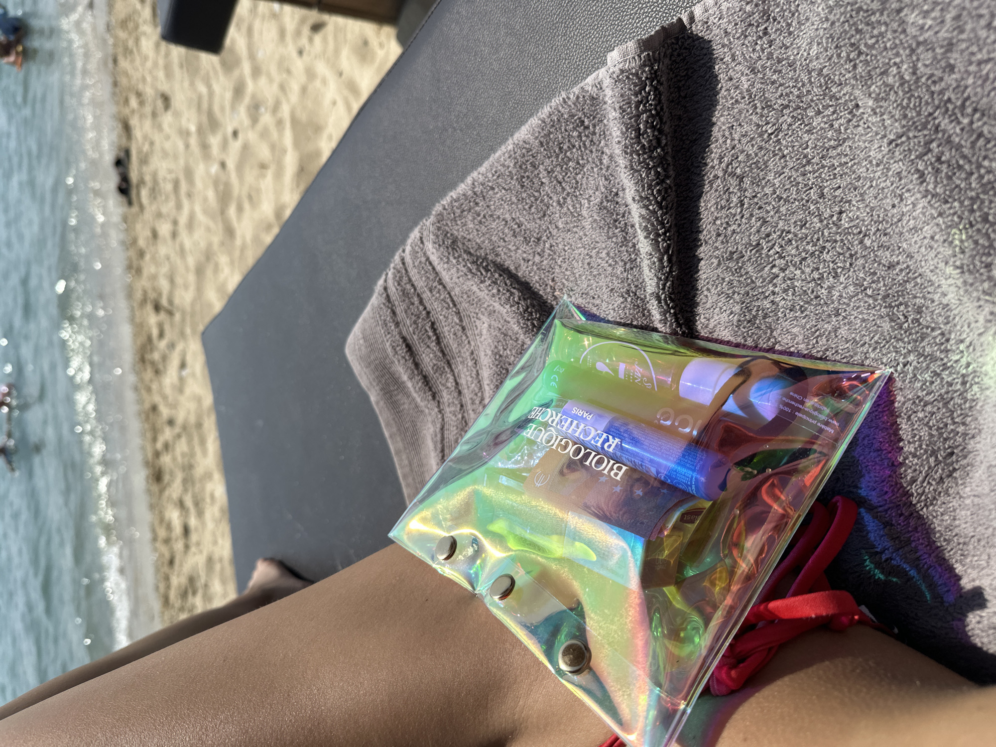 At Ofrynio Beach, Greece, a person is relaxing on a lounge chair with a towel. In the foreground, a clear, iridescent pouch filled with small beauty products is placed on the towel. One of the products is labeled "BIOLOGIQUE RECHERCHE PARIS". The person's legs and a red bikini strap are visible. In the background, the sea with gentle waves and a sandy beach can be seen.