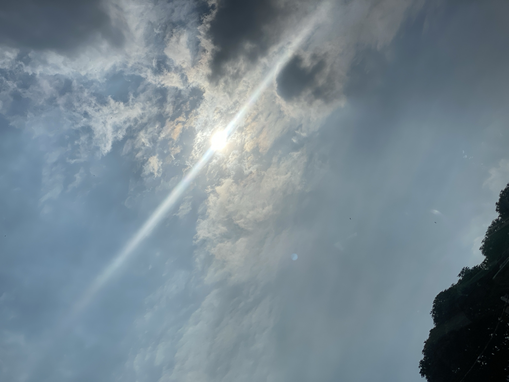 The image shows a sky with scattered clouds and a bright sunbeam cutting diagonally across it. The sun itself is partially obscured by clouds, creating a lens flare effect. In the lower portion of the image, the dark silhouette of trees is visible against the sky. There are no people or specific objects or actions depicted in the scene. The location is identified as Gambut Barat, Indonesia.