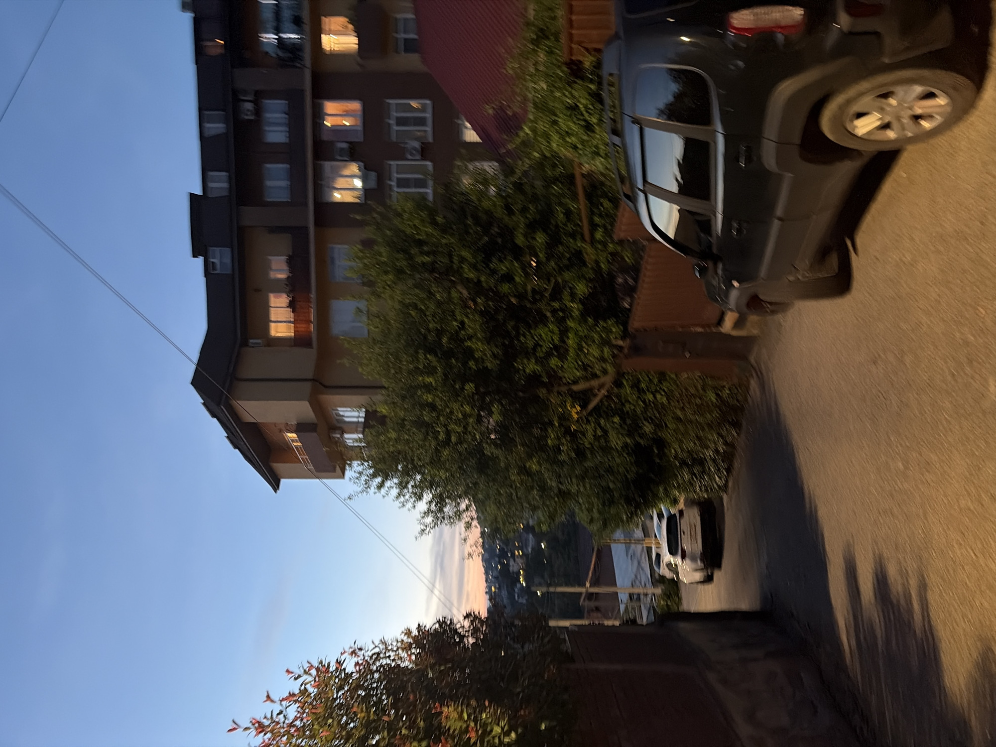 A street scene in Sochi, Russia, during dusk. The sky is a deep blue with streaks of orange and pink on the horizon, indicating sunset. A multi-story apartment building with illuminated windows stands on the right side of the street, its facade a mixture of beige and darker brown. Several trees with dense green foliage are situated in front of the building. On the left side of the street, a brick wall runs along the side of the road, with a tree bearing red leaves.
The street itself is paved and slopes downwards. Two cars are visible: a white hatchback parked further up the street, and a dark-colored SUV in the foreground, partially obscuring the view of the white car. The SUV is parked on the right side of the road, facing away from the viewer. The image has a slightly blurred quality, suggesting it was taken while in motion or with a fast shutter speed.